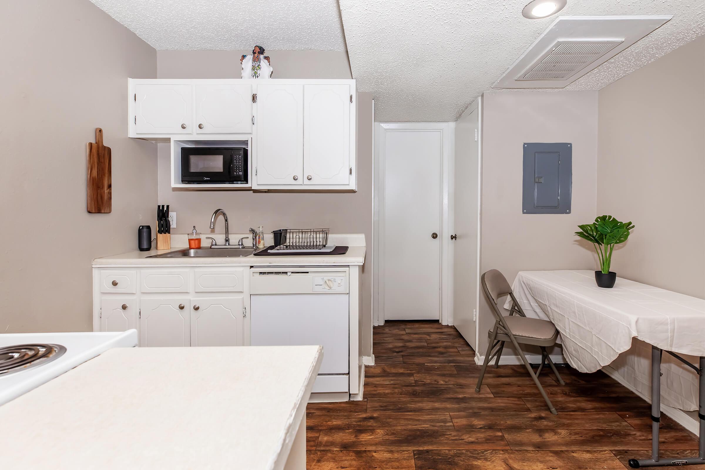 A small kitchen featuring white cabinets and appliances, including a microwave and a dishwasher. The countertop is clean and uncluttered, with a small plant on the table. A folding chair is placed next to a table draped in a white tablecloth. The flooring is wooden, and there is a closed door leading to another room.