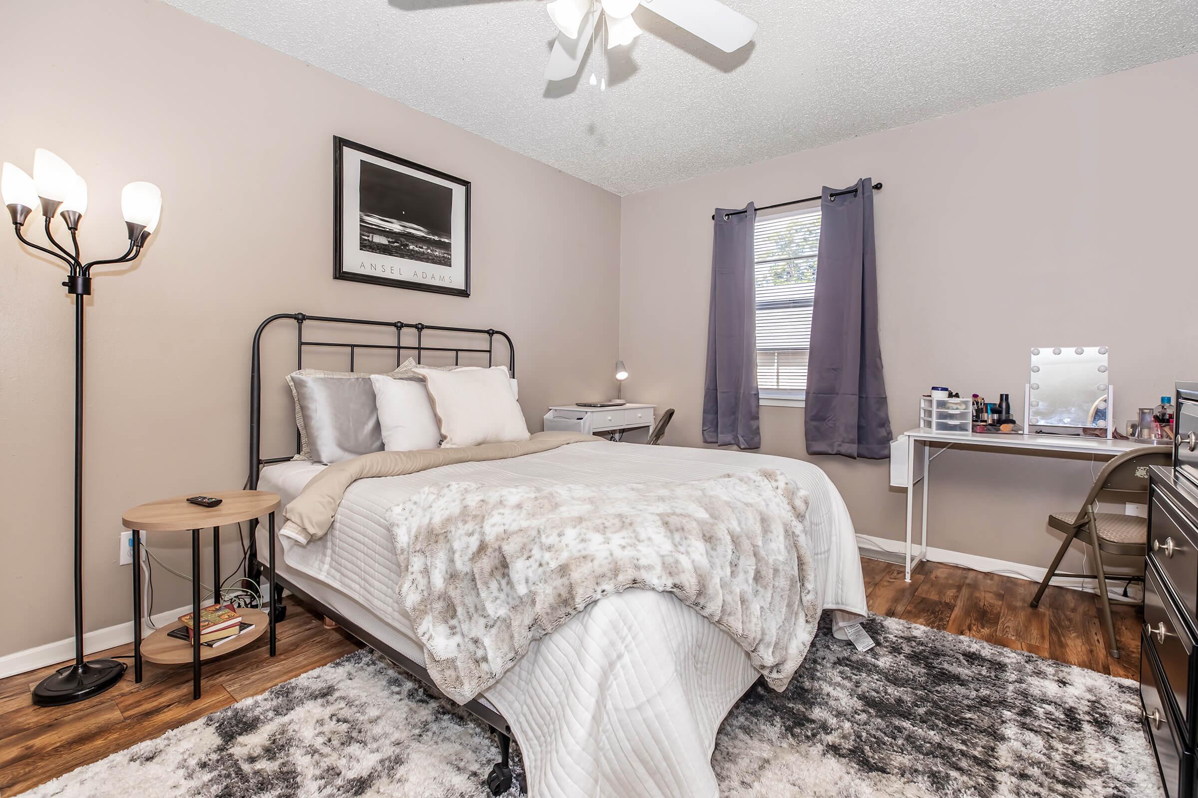 A cozy bedroom featuring a bed with a gray and white blanket, a wooden bedside table with a lamp, and a makeup vanity with a mirror. There are two windows with light curtains allowing natural light, and a soft area rug covers the floor. A ceiling fan provides ventilation. The walls are painted a warm neutral color.