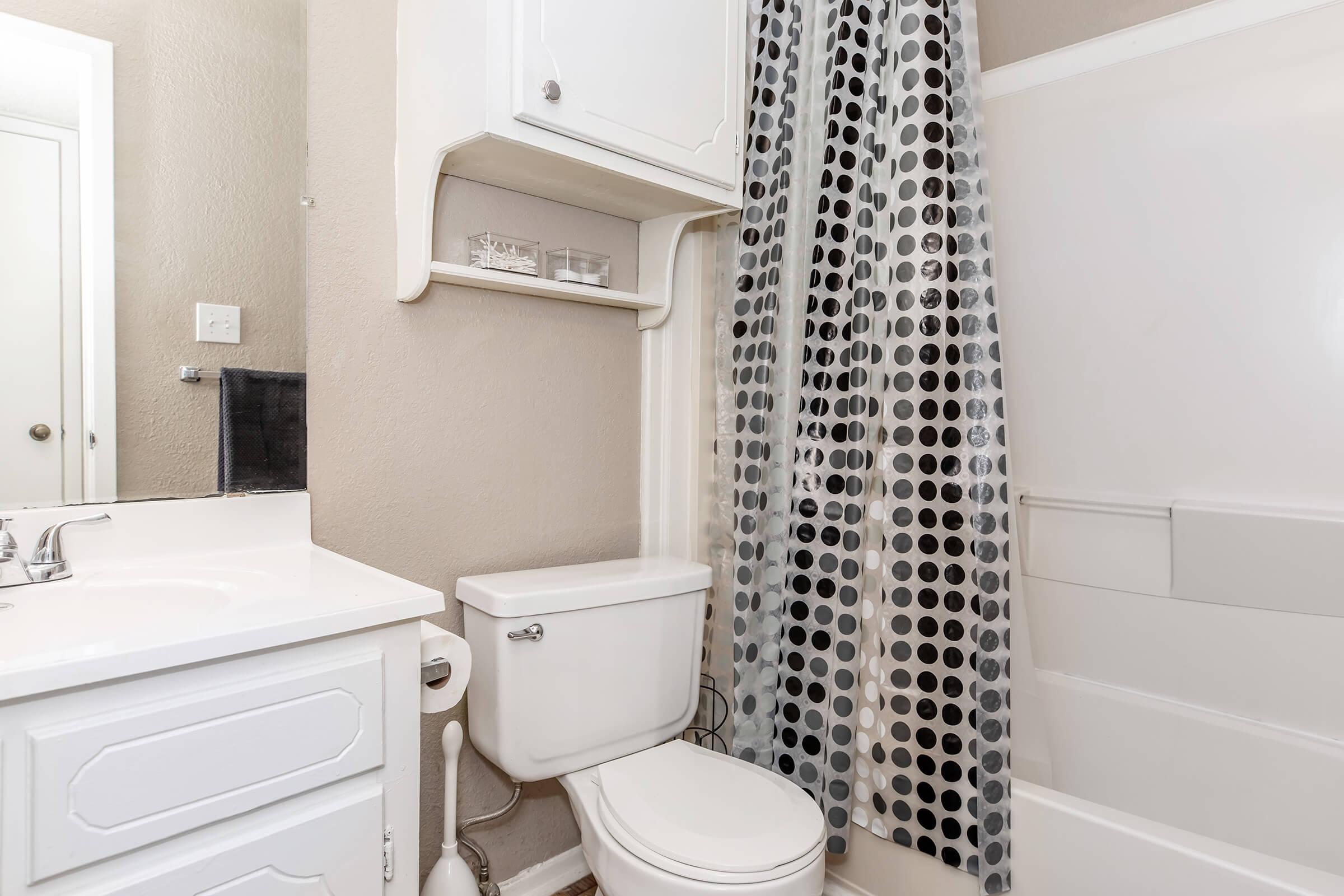 A small bathroom featuring a white toilet, sink with cabinet, and a shower with a polka dot curtain. The walls are painted light, and there is a shelf above the toilet that holds decorative items. The space is well-lit and tidy, with a minimalist design.