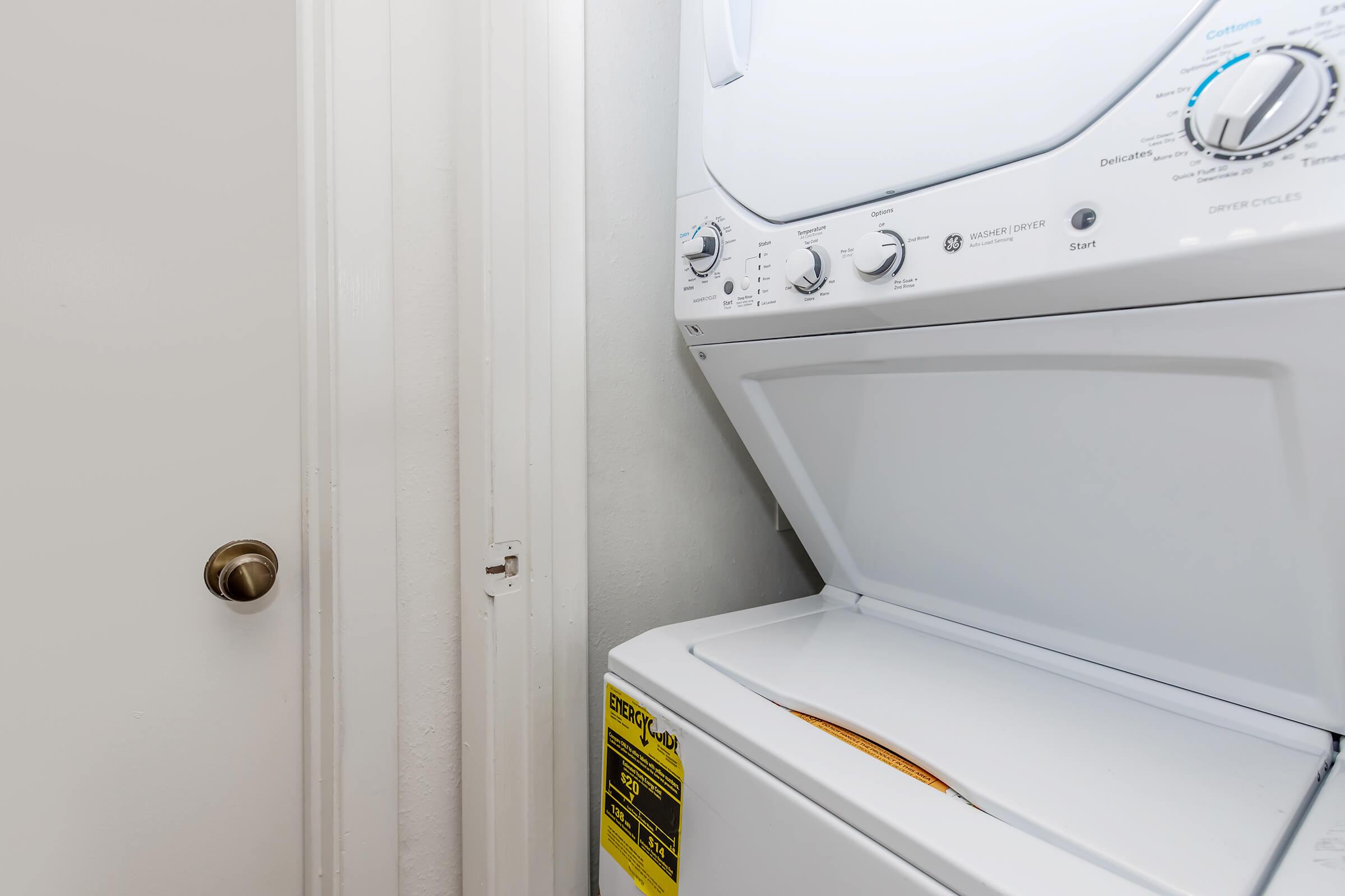 A stacked washer and dryer located in a small laundry space. The dryer is on top, featuring control knobs and a start button. The wall beside the appliances is plain, and there is a doorknob on the adjacent wall, suggesting proximity to a door. The overall setting is compact and functional.