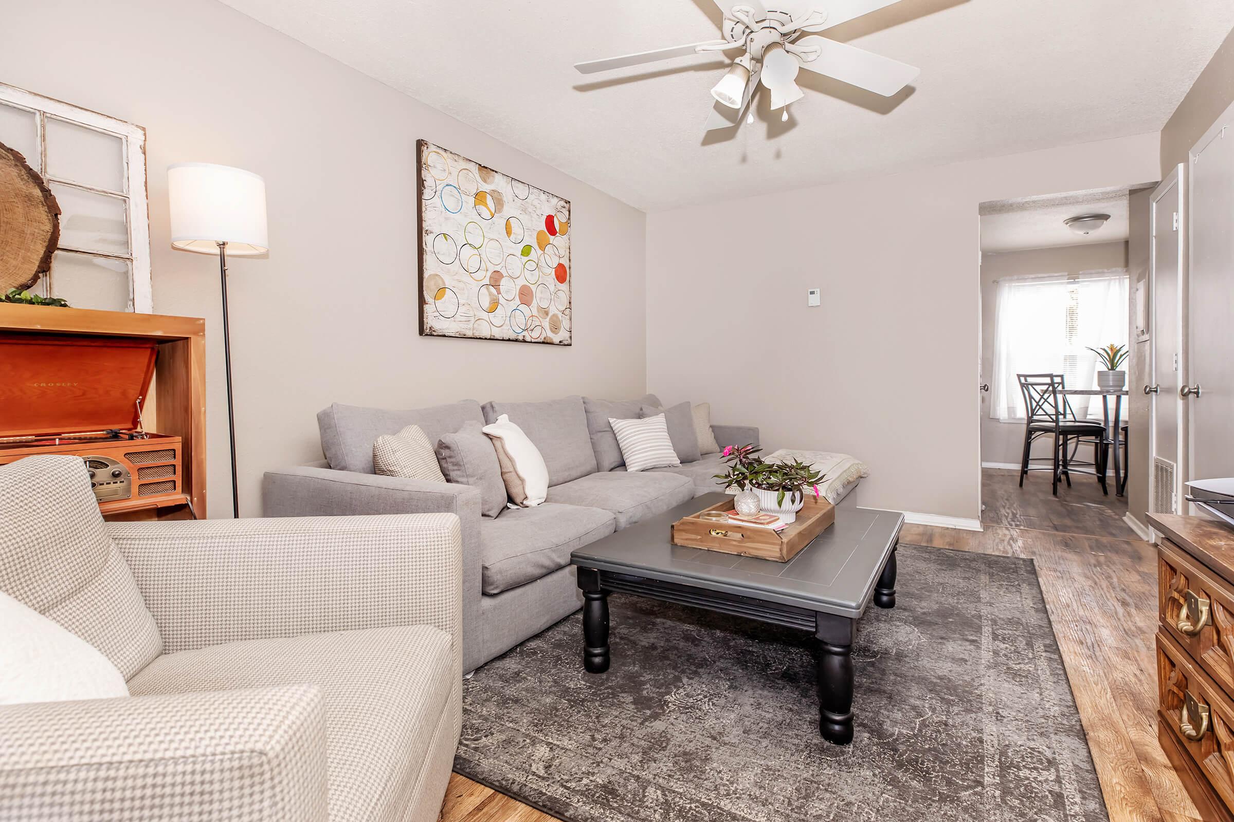 A cozy living room featuring a light gray sofa with decorative pillows, a dark coffee table, and a patterned rug. There's a wooden side table with a lamp and a piece of wall art above the couch. In the background, a doorway leads to a bright dining area. A ceiling fan adds a touch of comfort.