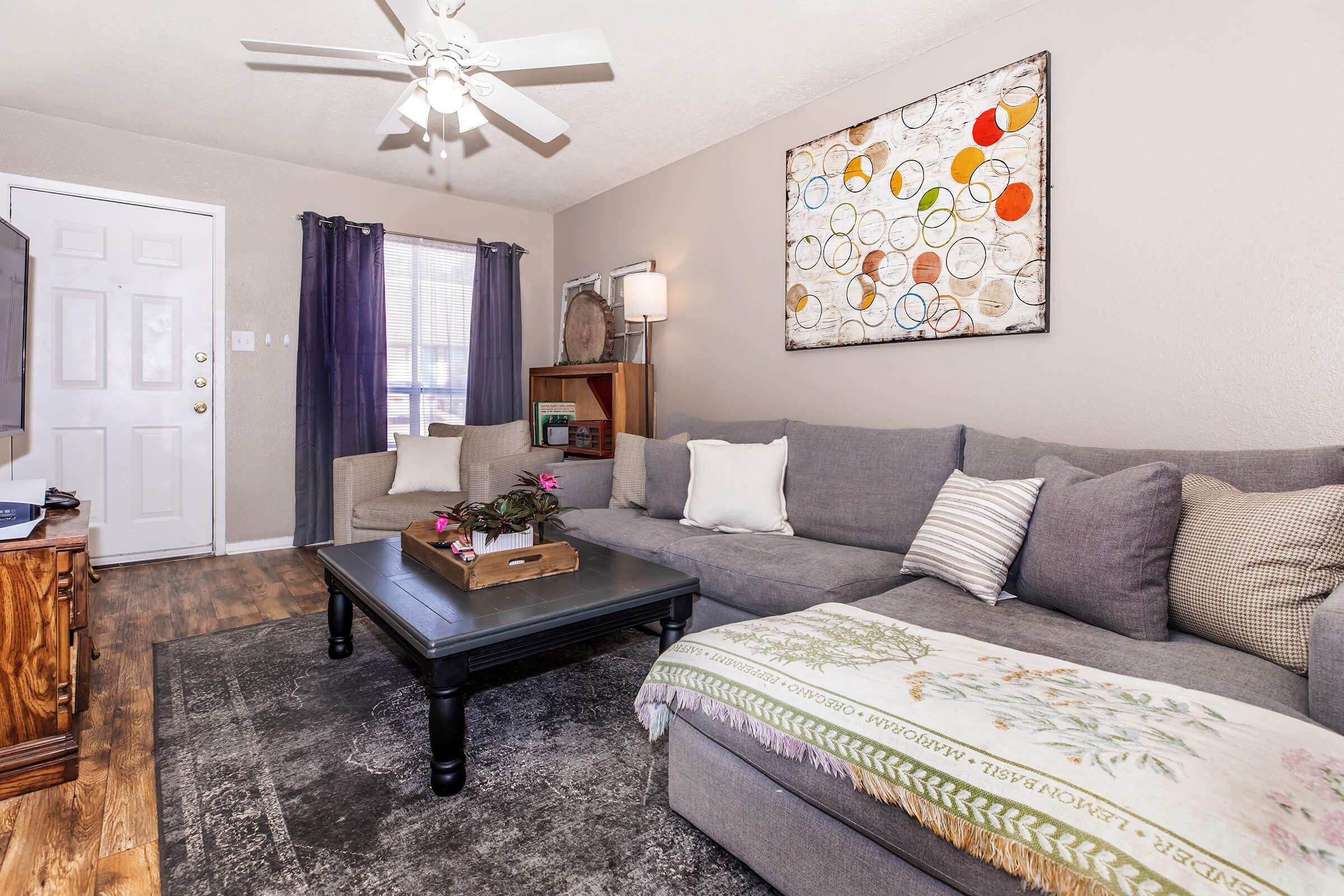 A cozy living room featuring a gray sectional sofa with decorative pillows, an accent table with a small plant, and a textured area rug. A large piece of abstract wall art hangs above the sofa, and a ceiling fan adds a touch of comfort. Light enters through a window with sheer curtains.