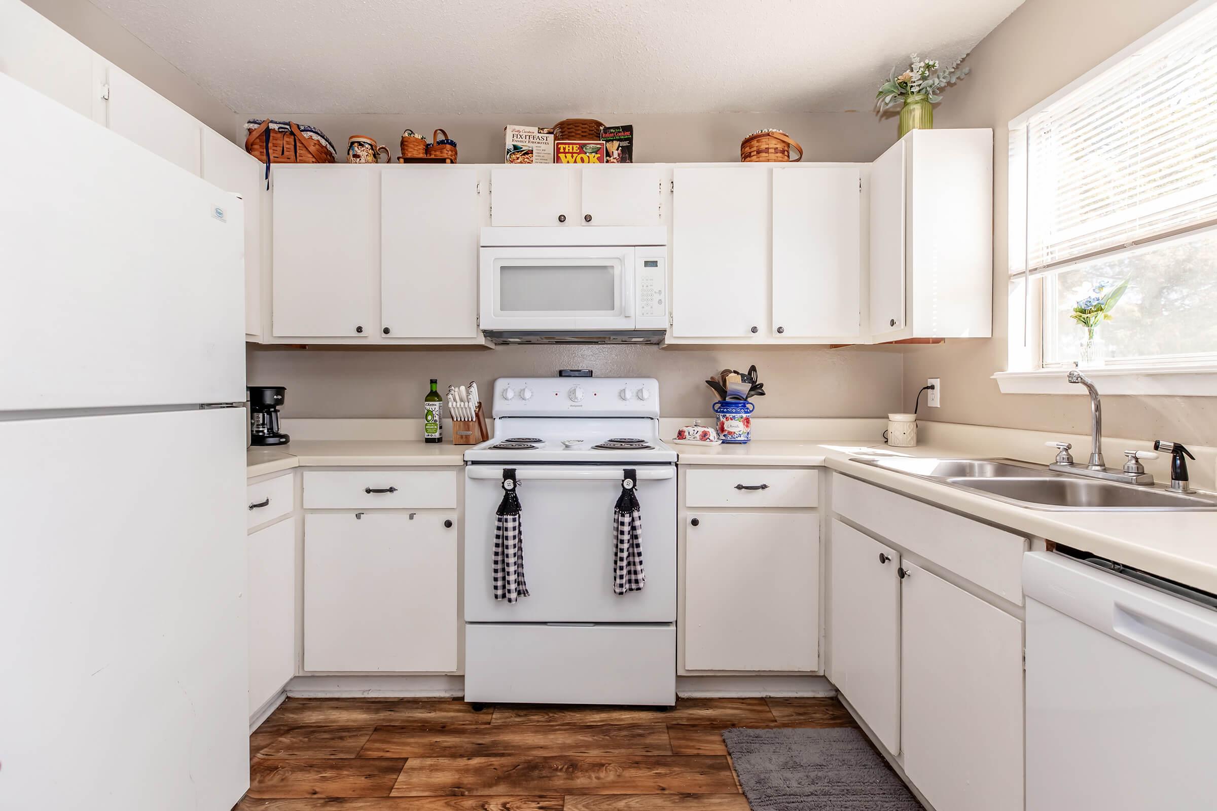 A bright kitchen featuring white cabinetry, a white refrigerator, and a stove with a microwave above. The countertop has a dish rack and various kitchen items. Windows allow natural light, and decorative baskets are placed on top of the cabinets. The floor has a wood-like appearance, enhancing the cozy atmosphere.