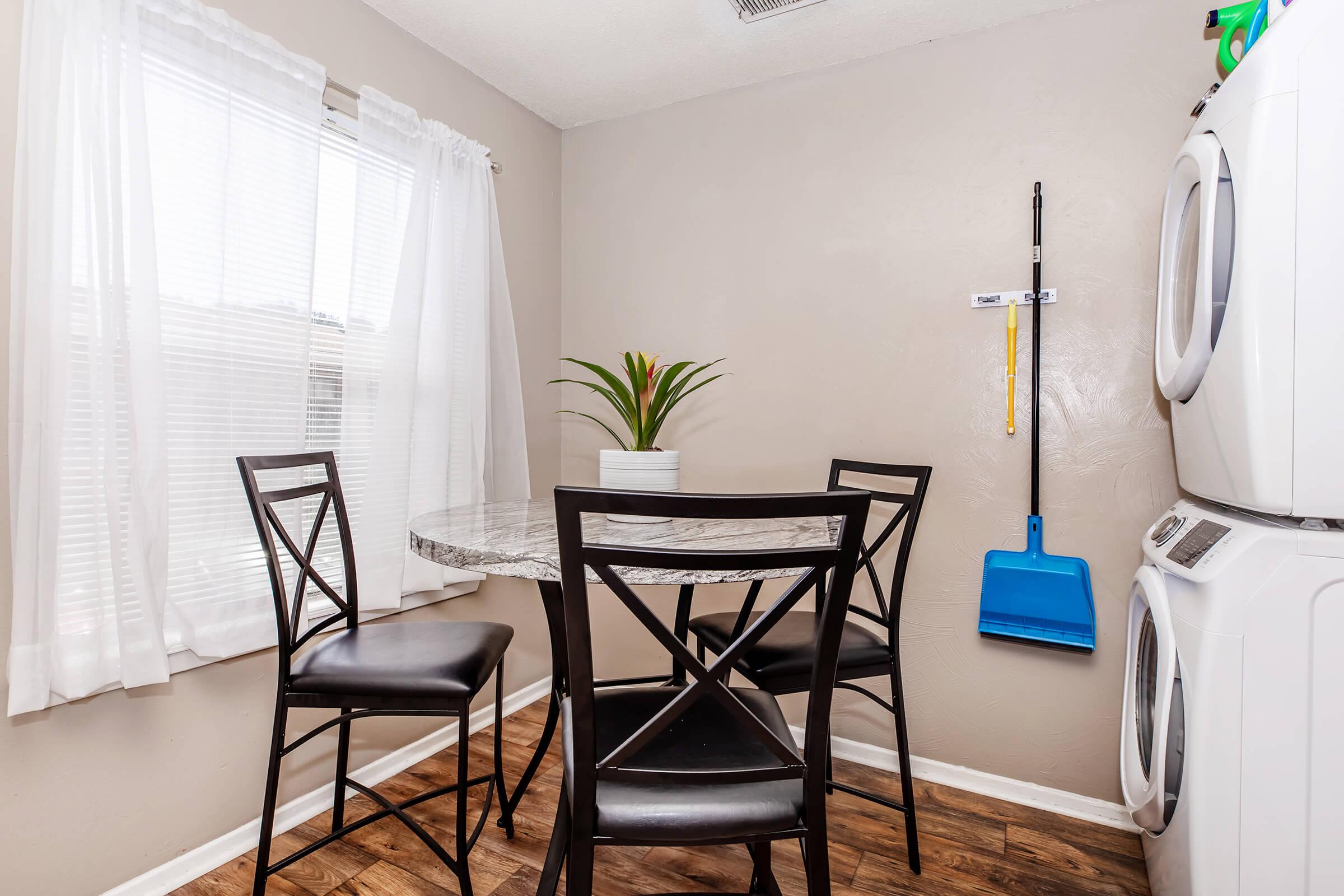 A small dining area featuring a round marble table with four black chairs. There’s a potted plant on the table, a window with sheer white curtains, and a wall-mounted blue dustpan next to a stacked washer and dryer. The flooring is wood-like laminate.