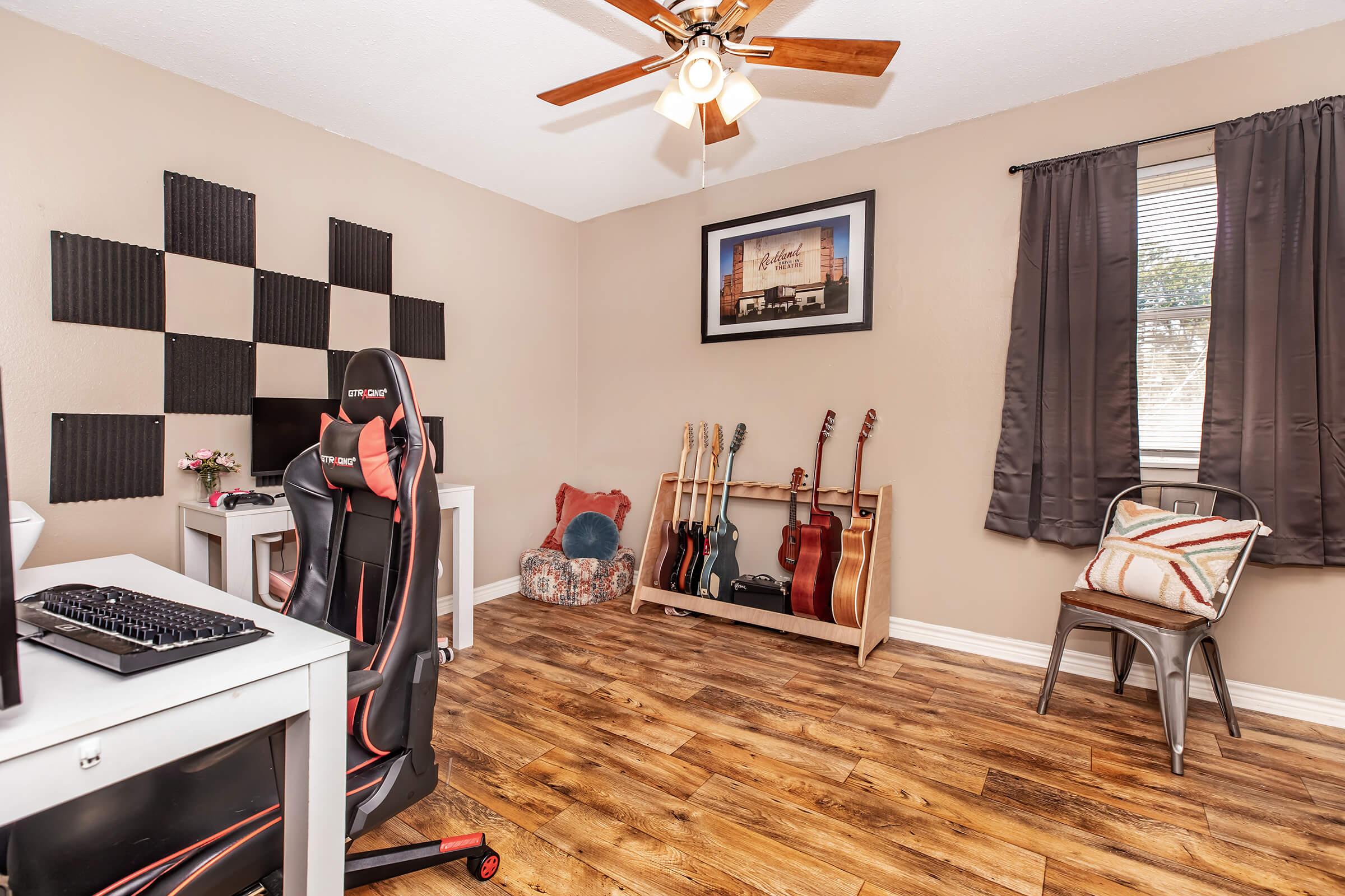 A cozy room featuring a home office setup with a desk and gaming chair, a wall decorated with black acoustic panels, and a corner displaying several guitars. Natural light filters through a window with dark curtains, complemented by a seating area with a patterned cushion and a simple metal chair.