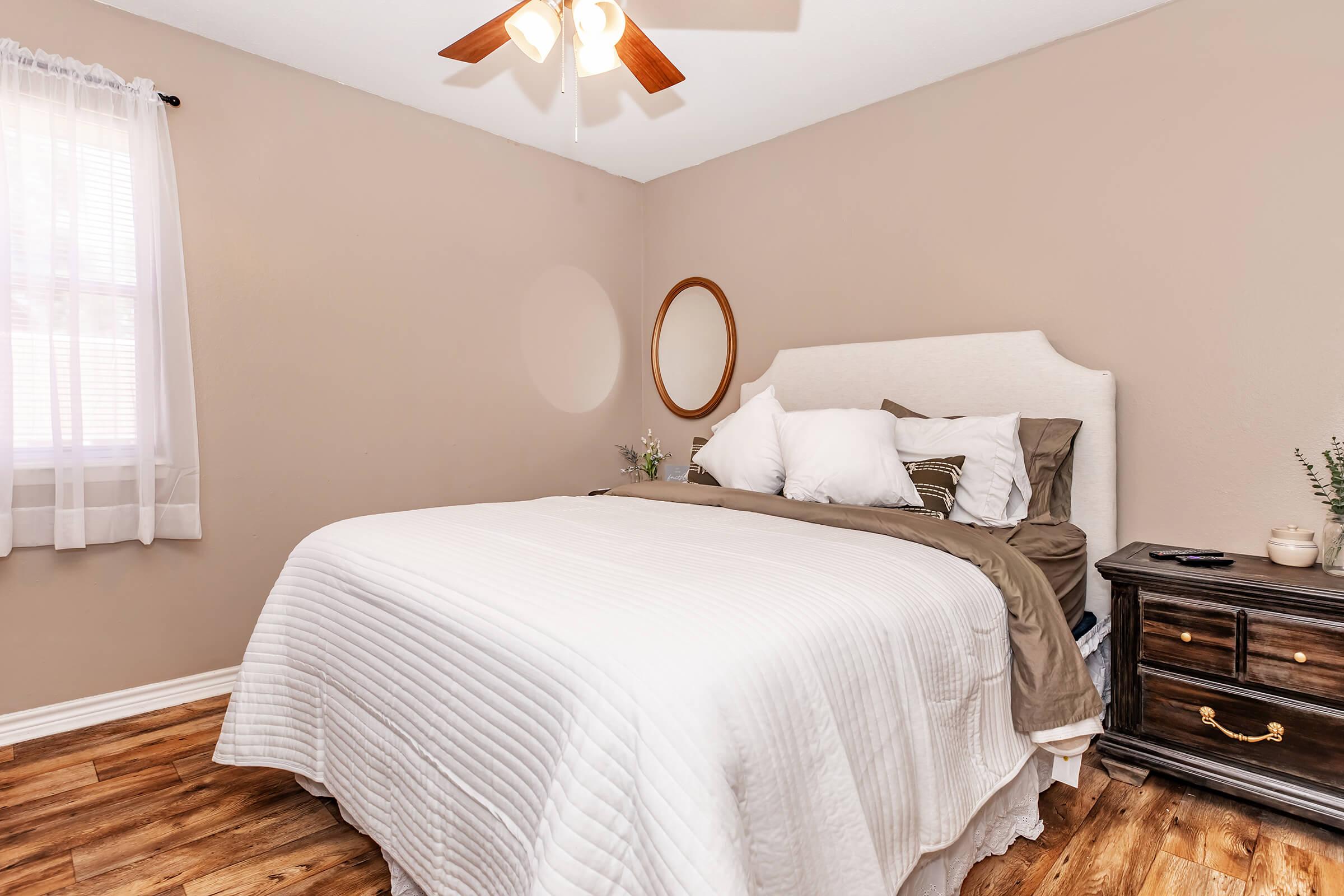 Cozy bedroom featuring a plush bed with white bedding, accented by several decorative pillows. The room has a ceiling fan, a round mirror above the headboard, and a dark wooden nightstand beside the bed. Natural light filters in through a window with sheer curtains, illuminating the warm, neutral-toned walls and wooden flooring.