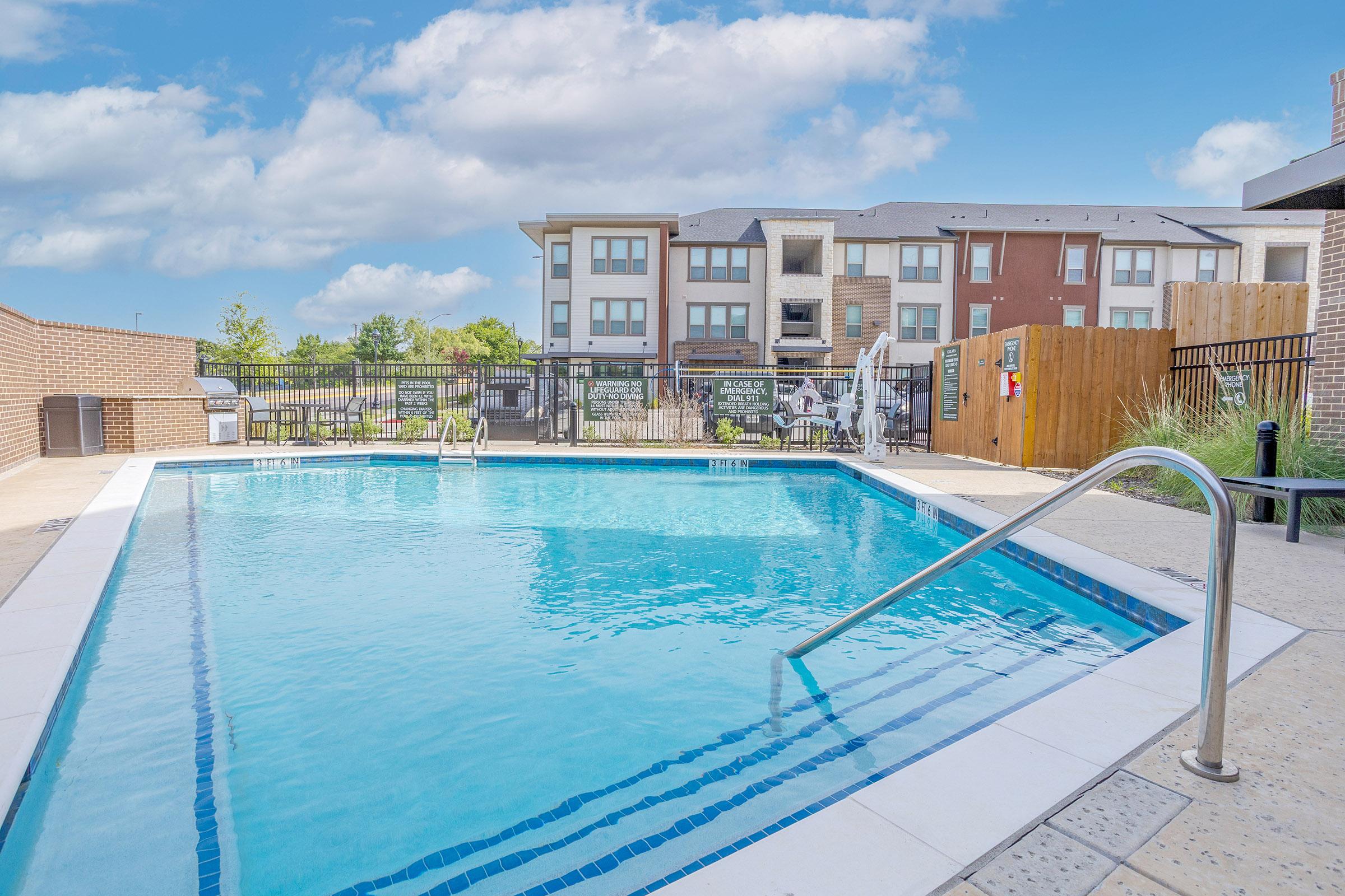 A clear blue swimming pool surrounded by a patio area, with lounge chairs and a fence in the background. The pool features a metal ladder for access, and modern apartment buildings are visible in the distance against a partly cloudy sky.