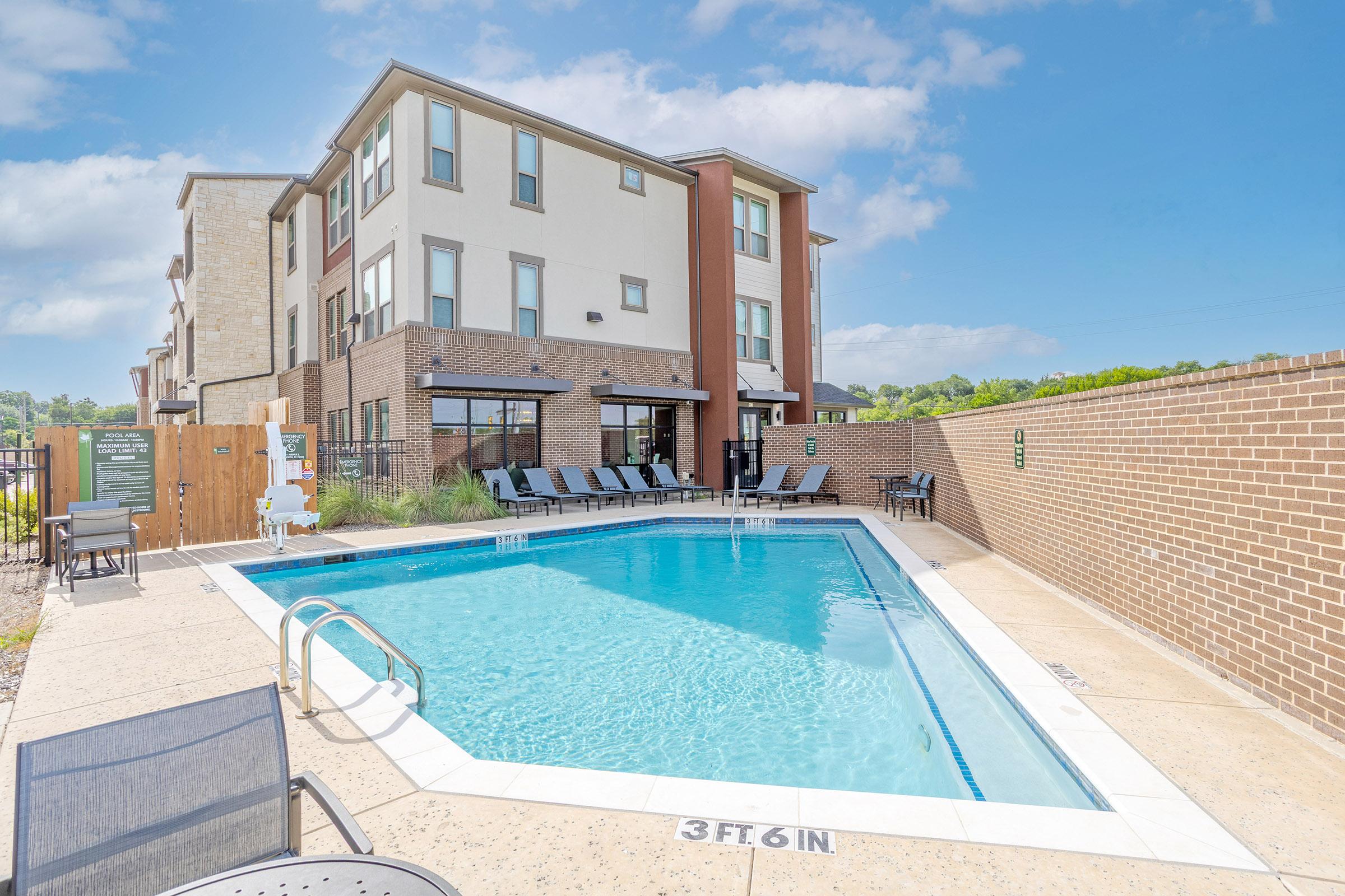 A clear blue swimming pool surrounded by lounge chairs and a brick wall, with a modern three-story building in the background. The area is well-lit under a sunny sky, creating a welcoming atmosphere for relaxation and leisure.