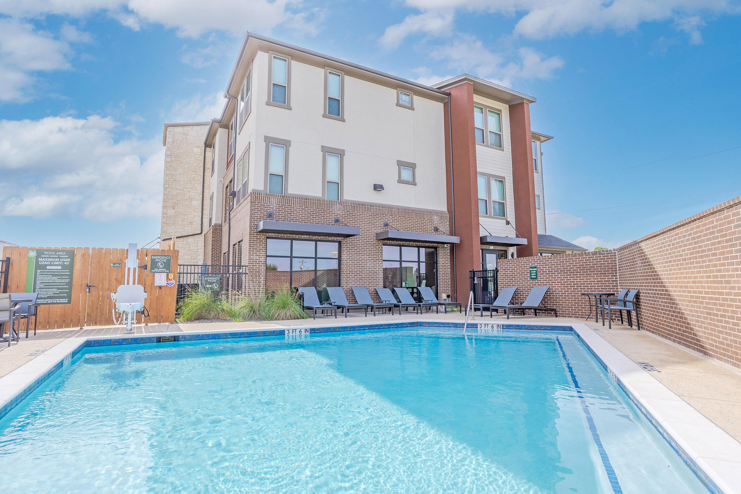 A modern apartment building with a swimming pool in the foreground. The pool features lounge chairs around it, while the building has large windows and a brick exterior. A wooden fence borders the area, and the sky is clear with a few clouds.