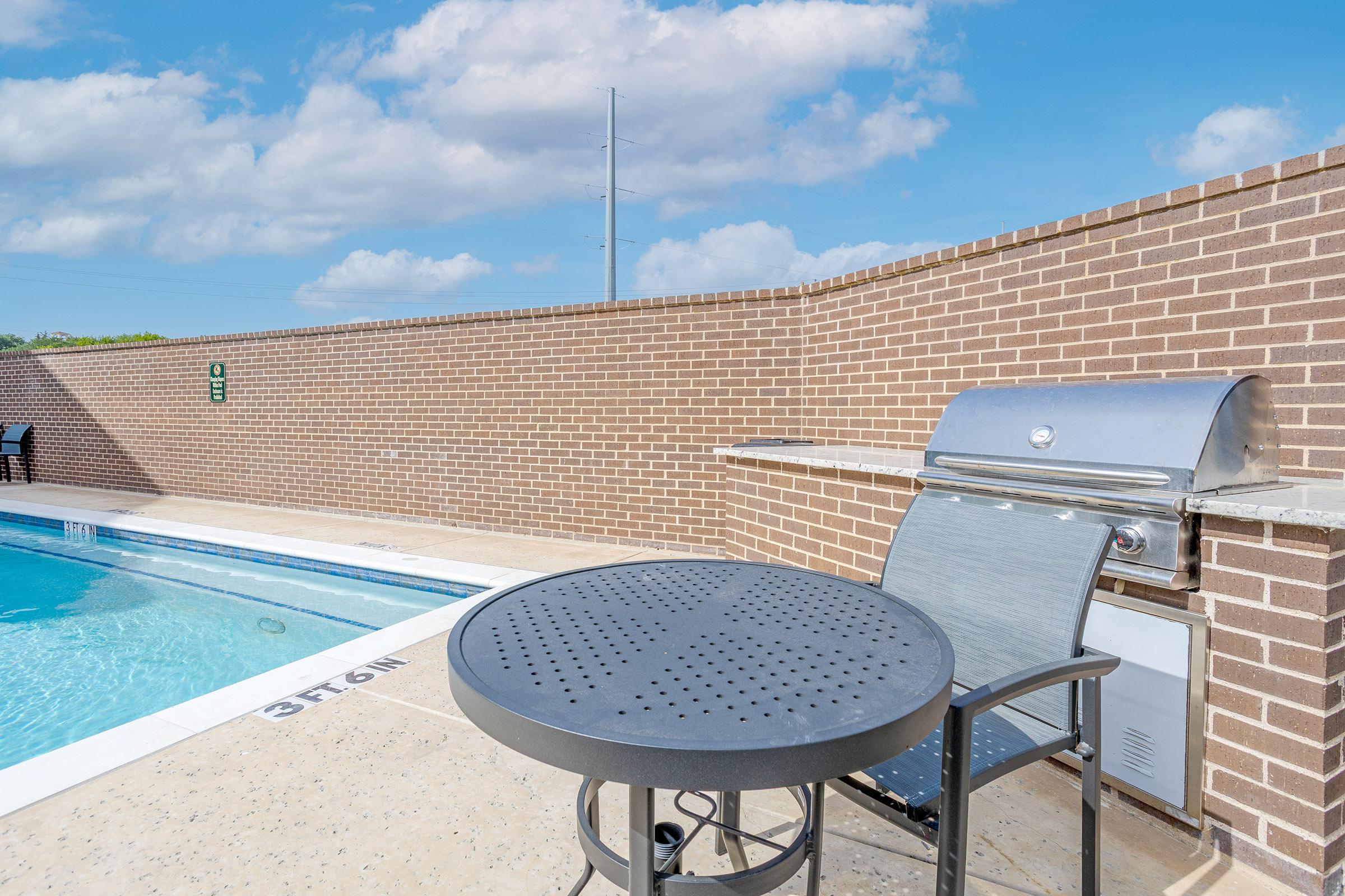A swimming pool with clear blue water, surrounded by a smooth deck. In the foreground, there is a round black table with a matching chair. Nearby, a stainless steel grill is built into a brick wall. The sky is partly cloudy, creating a bright and inviting atmosphere.
