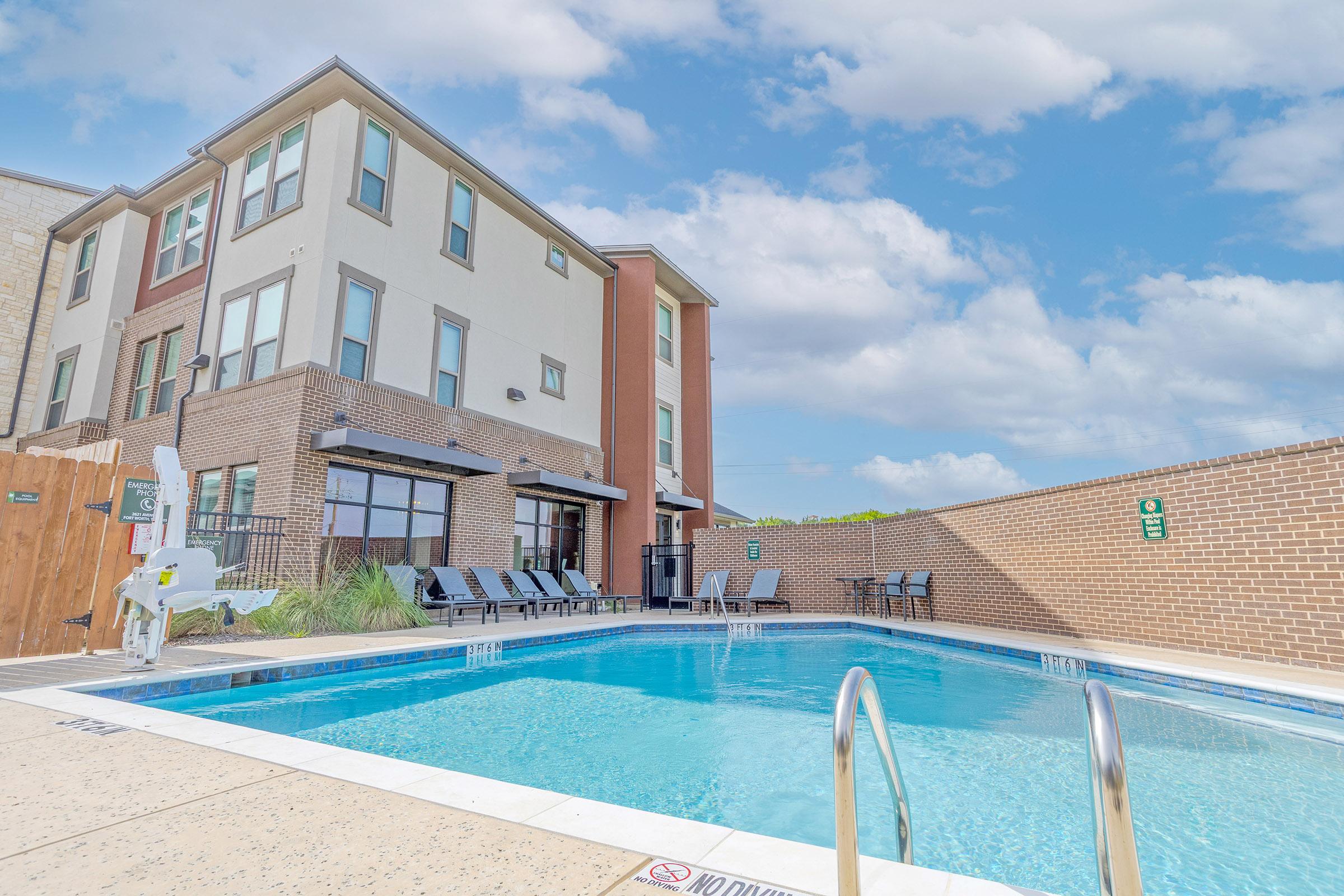 A sunny pool area featuring a clear blue swimming pool, surrounded by lounge chairs and a modern building in the background. The scene includes a well-maintained patio area with greenery and a clear sky showcasing fluffy white clouds.