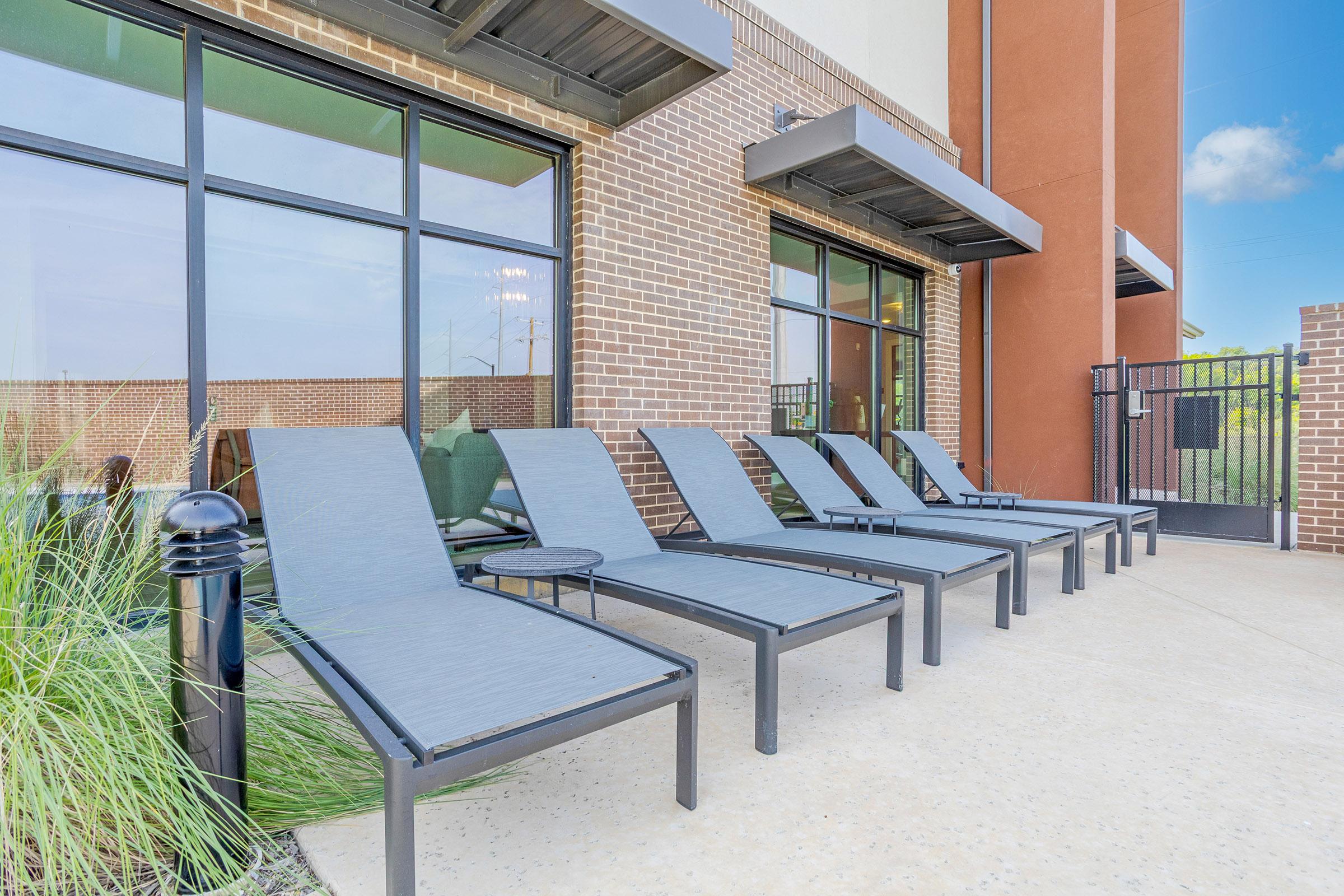 A row of modern poolside lounge chairs arranged neatly outside a building, with large windows visible behind them. The area features a paved surface and some greenery, creating a relaxed outdoor atmosphere.