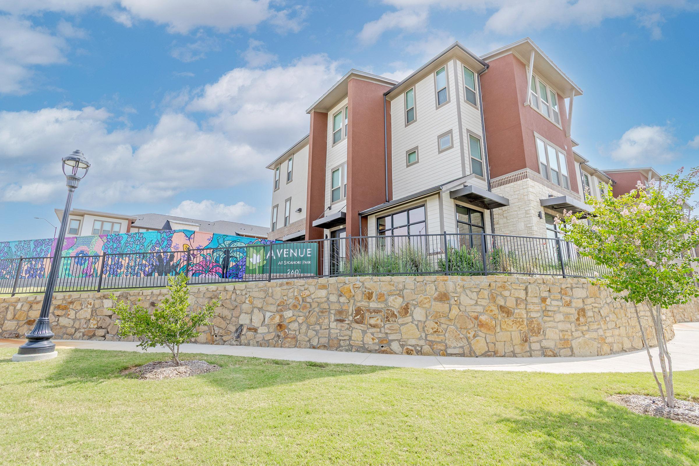A modern apartment building featuring a combination of red and beige siding, with a decorative mural on a fence nearby. The structure is set against a blue sky with scattered clouds, and there is a landscaped area in the foreground with green grass and trees. A lamp post is visible to the left.