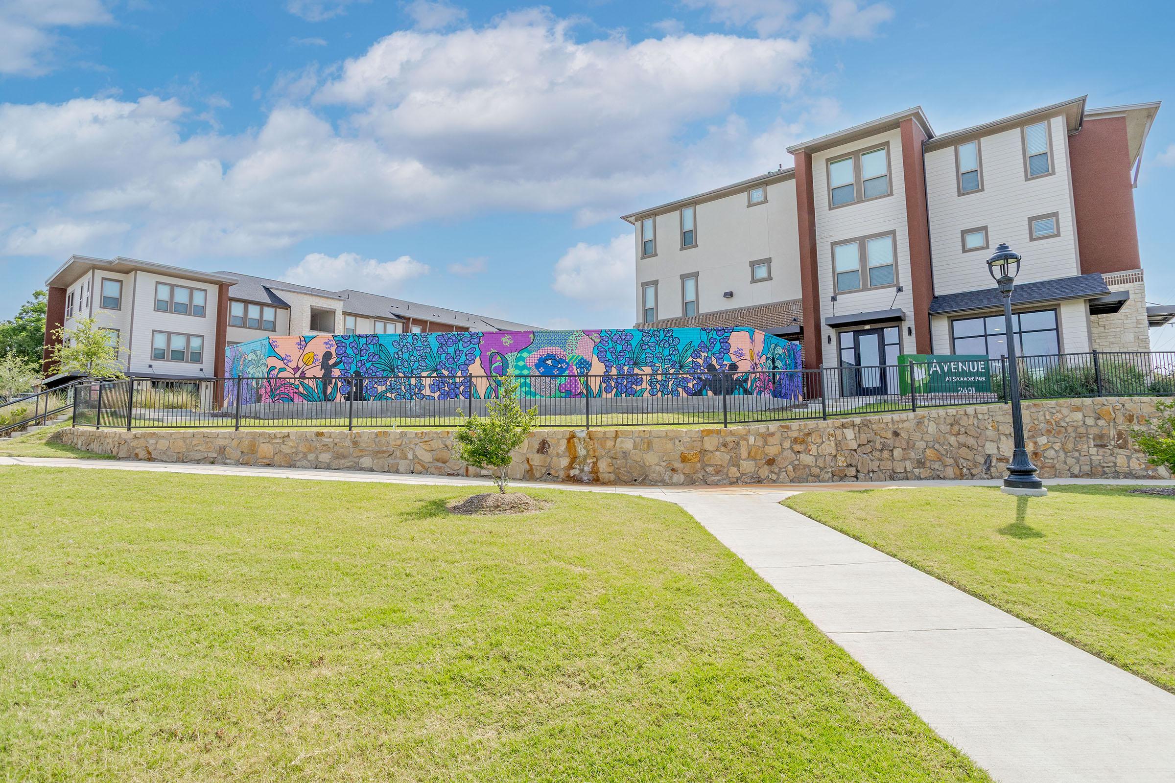 A colorful mural depicting vibrant flowers and abstract designs on a wall, surrounded by green grass. In the background, there are modern apartment buildings with a sign labeled "Avenue." The scene is under a partly cloudy sky, showcasing a community area with a walkway.