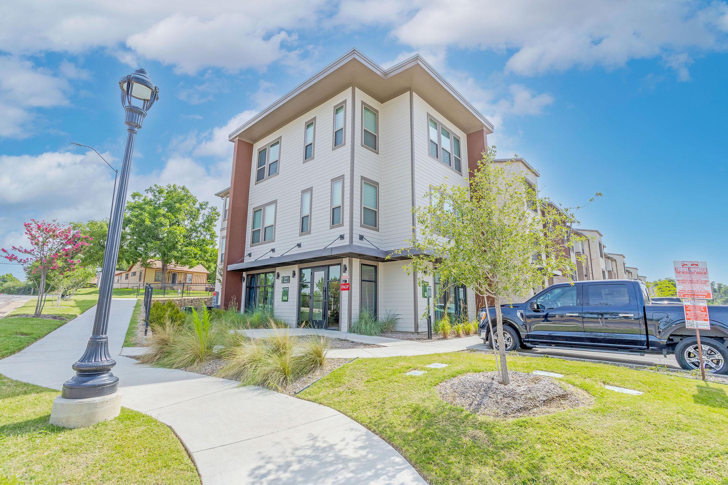 A modern three-story apartment building with a mix of light and dark siding. It features large windows and a landscaped front area with trees and grass. A black pickup truck is parked nearby, and a lamppost adds to the outdoor ambiance under a bright blue sky with a few clouds.