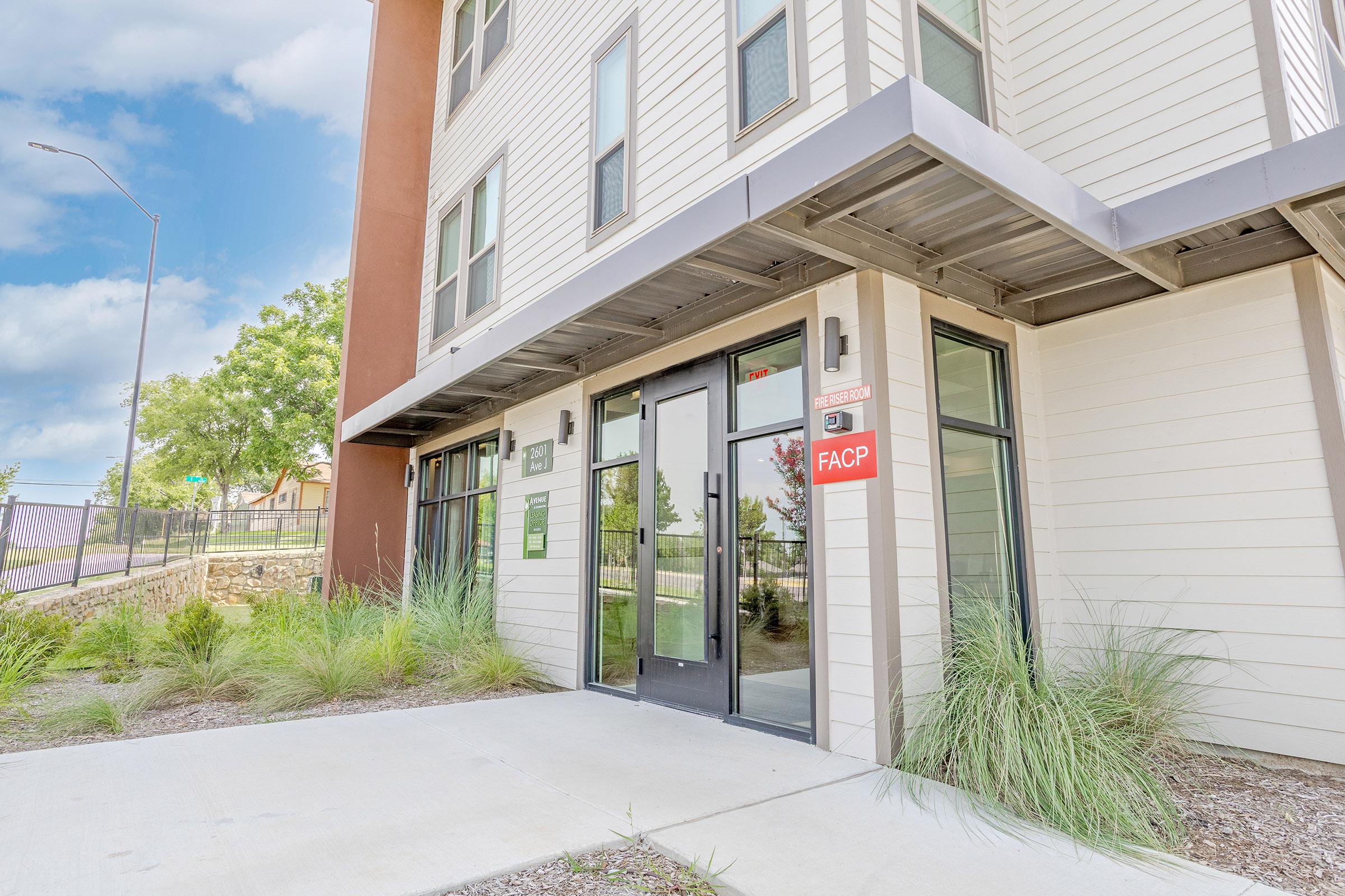 Modern building entrance featuring large glass doors, a sign labeled "FACP," and landscaped surroundings with greenery. The structure has a contemporary design with a mix of materials and colors, situated in a well-lit area with blue skies and clouds in the background.
