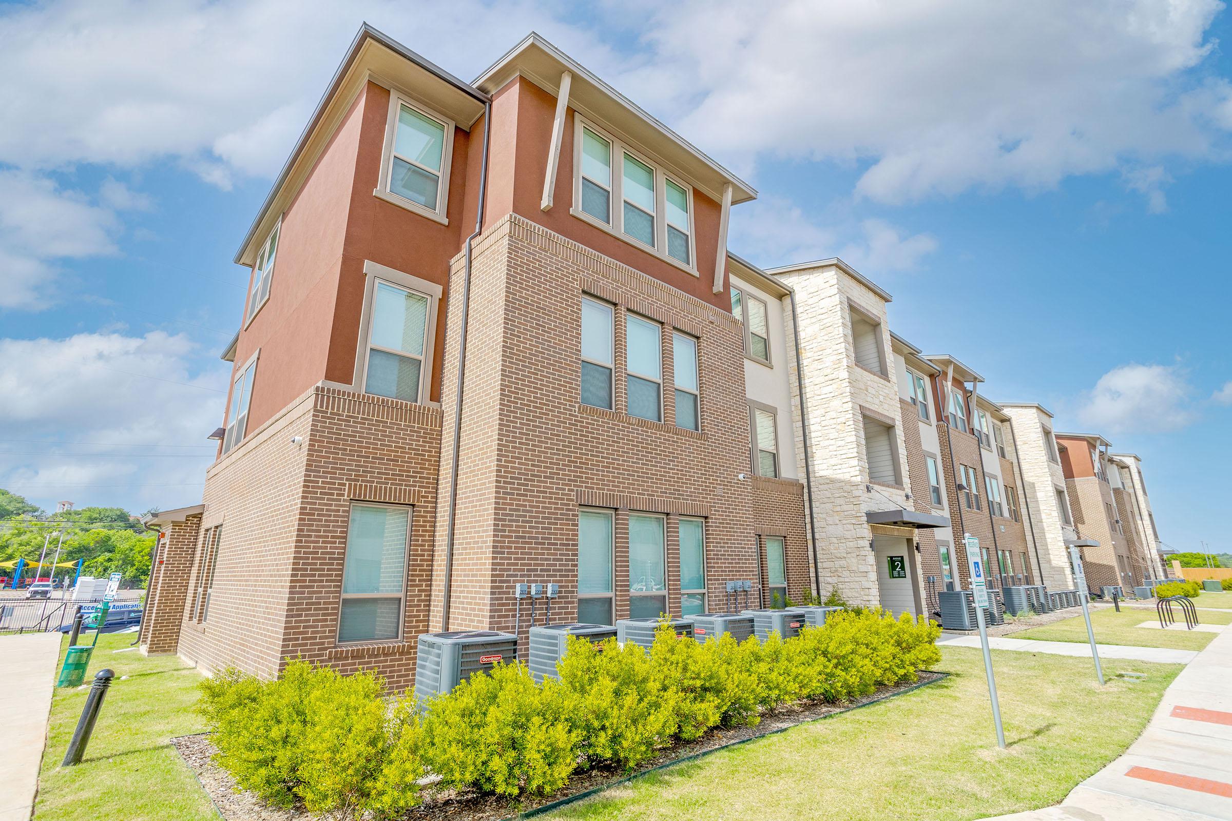 Modern multi-story apartment building with brick and stone façade, featuring large windows and air conditioning units outside. The surrounding area is landscaped with green shrubs under a clear blue sky with scattered clouds.