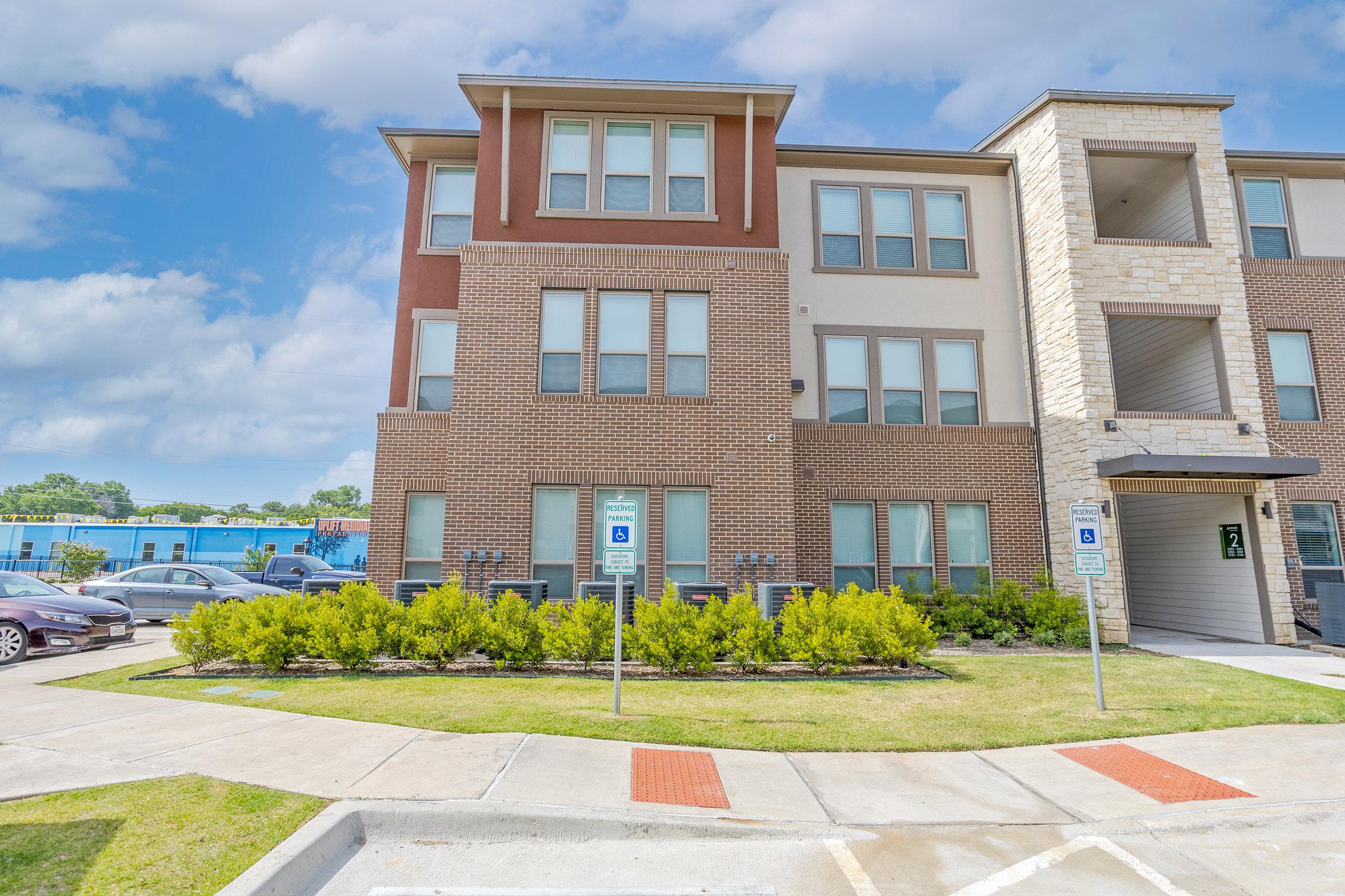 Modern multi-family apartment building with a mix of brick and stucco exteriors. The front features large windows and a covered entryway. Accessible parking spaces are available in front of the building, and landscaping includes neatly trimmed hedges and a grassy area. Clear blue sky adds a bright atmosphere.