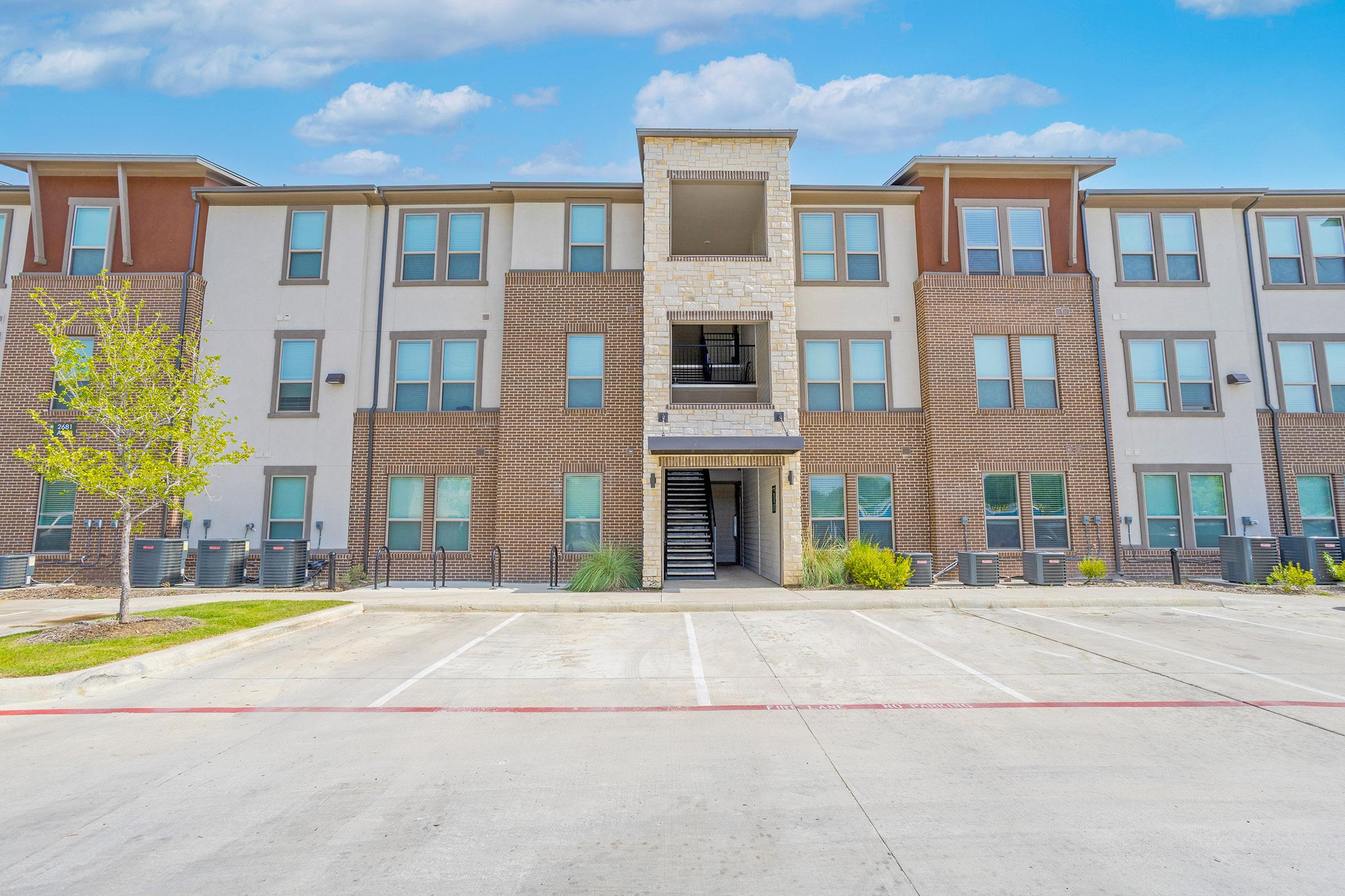 A modern apartment complex featuring three stories of brick and stone architecture. The center has a prominent entrance with stairs leading up, flanked by large windows. Nearby are small trees and shrubs, with a concrete parking area in front and a clear blue sky overhead.