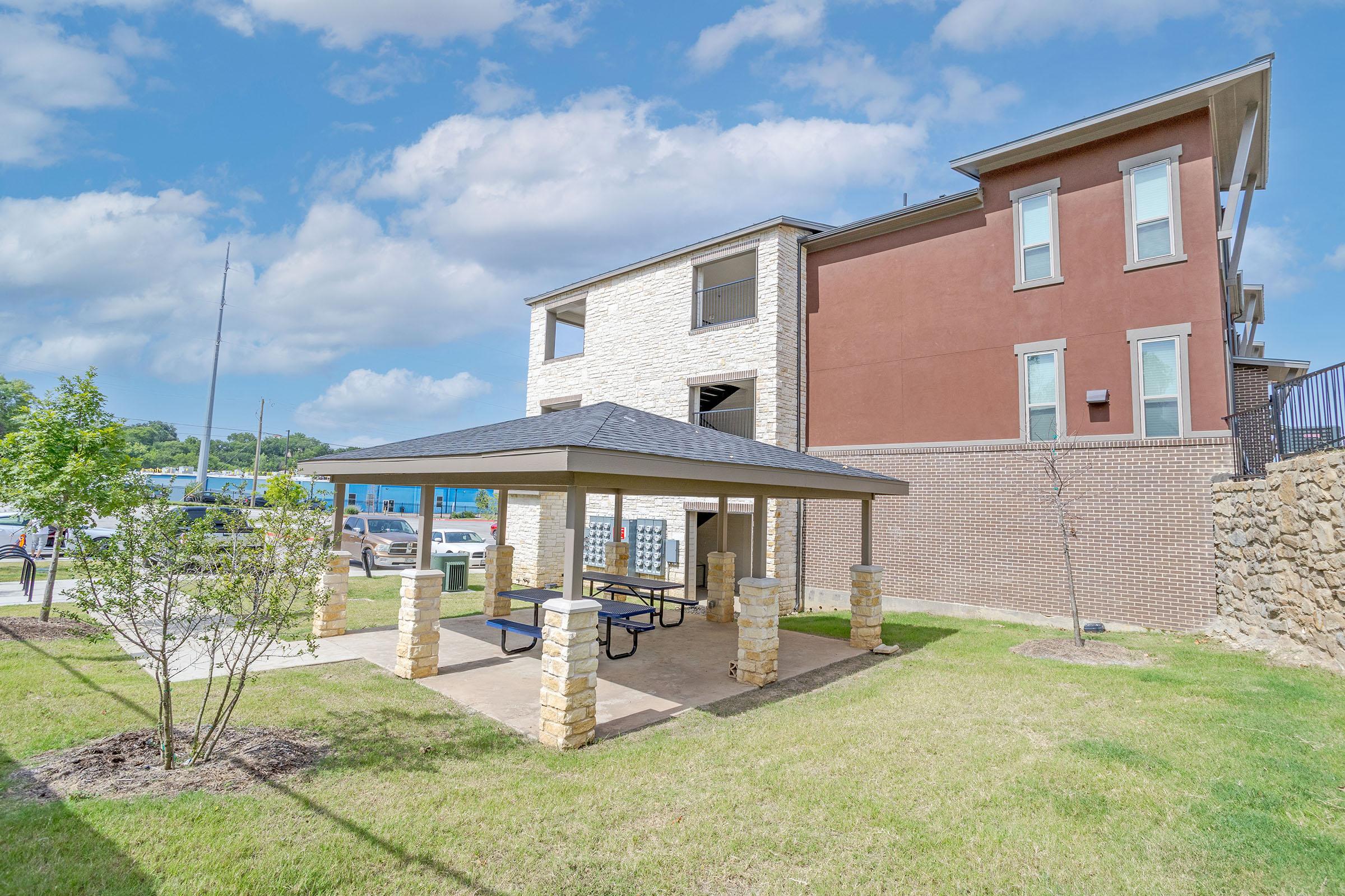 A view of a landscaped outdoor area featuring a covered picnic shelter with tables, surrounded by grass and small trees. In the background, there are modern apartment buildings with a mix of brick and stone exteriors, under a partly cloudy sky. Vehicles are parked nearby.