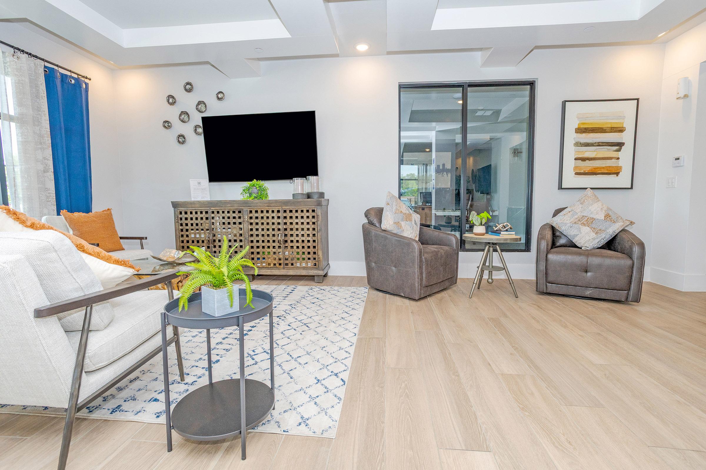 A modern living room featuring two grey armchairs and a white sofa. A round side table holds a small plant. The room includes a large TV on a cabinet, a textured blue and white rug, and a decorative wall mirror. Natural light comes through large windows with sheer curtains.