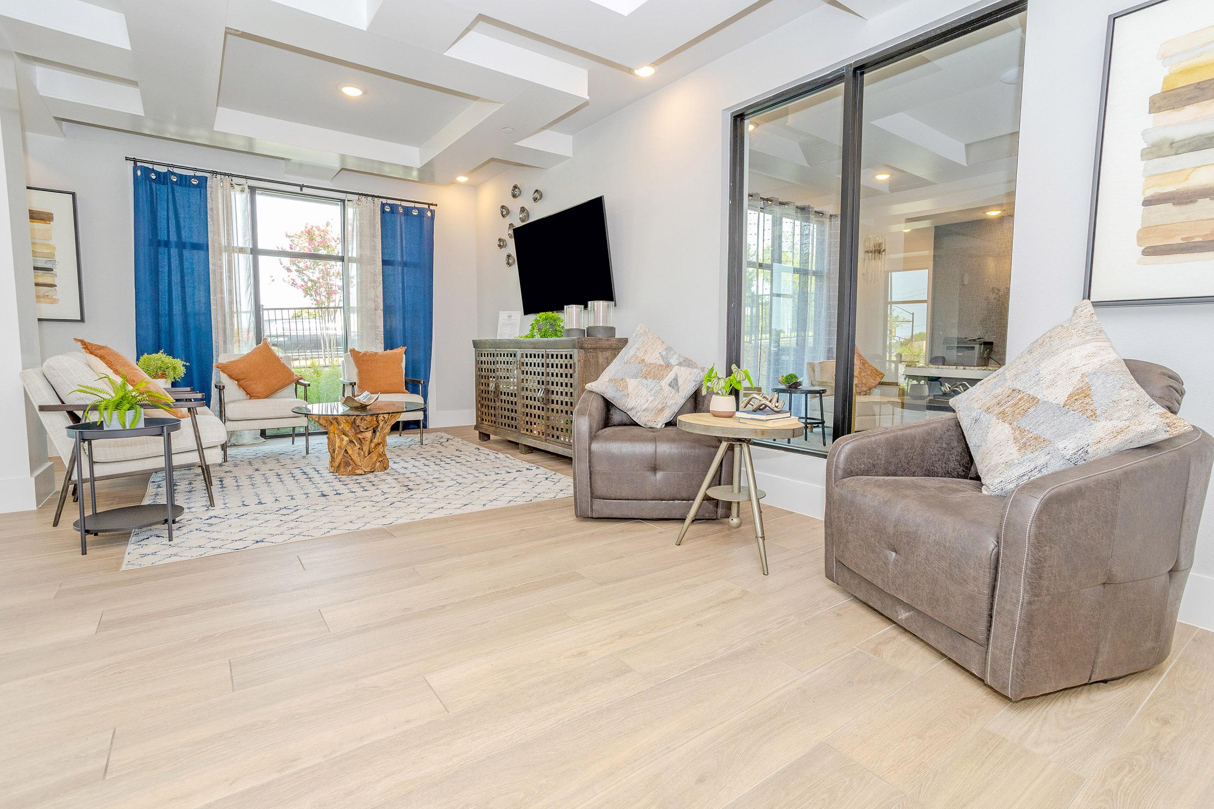 A modern living room featuring a light-colored wooden floor, two comfortable chairs, a coffee table, and a decorative rug. The space is enhanced by blue curtains, green plants, and cushions, with a large window and a TV on the wall, creating a bright and inviting atmosphere.