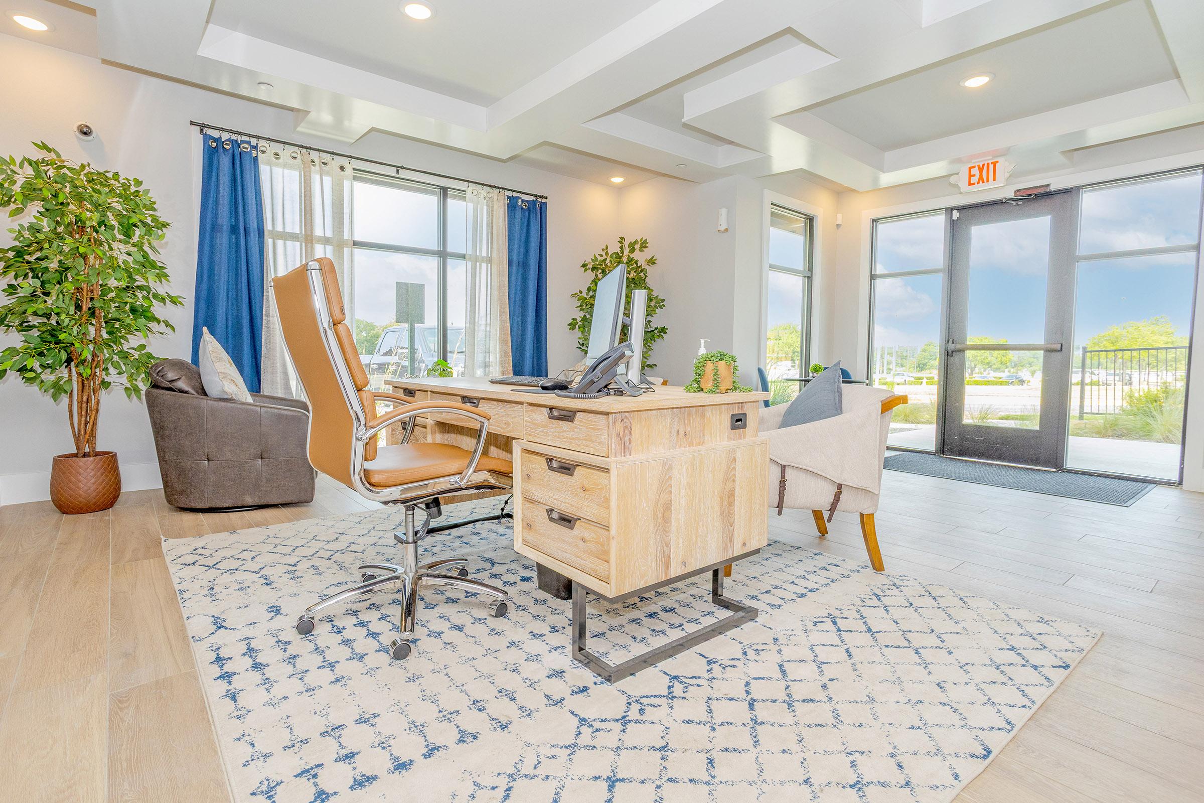 A modern office space featuring a wooden desk with a phone and decorative plants. There is a comfortable chair, a sofa, and a large window with blue curtains allowing natural light to fill the room. The floor has light wood planks, and a patterned area rug adds warmth to the space. A door leads outside.