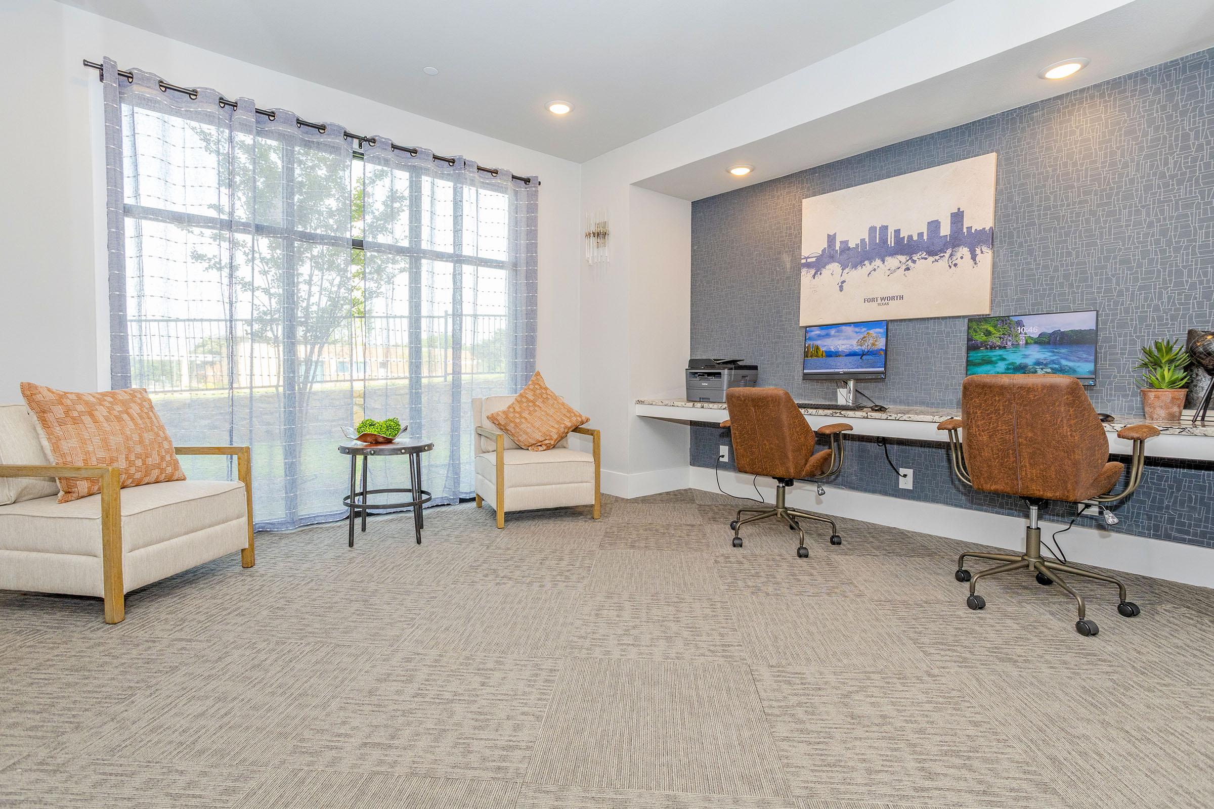 A cozy office space featuring two chairs with orange accent pillows, a small table with a plant, and a work desk with two computer monitors. Natural light streams in through large windows, with a view of greenery outside. The walls are decorated with a cityscape art piece and a textured blue accent wall.