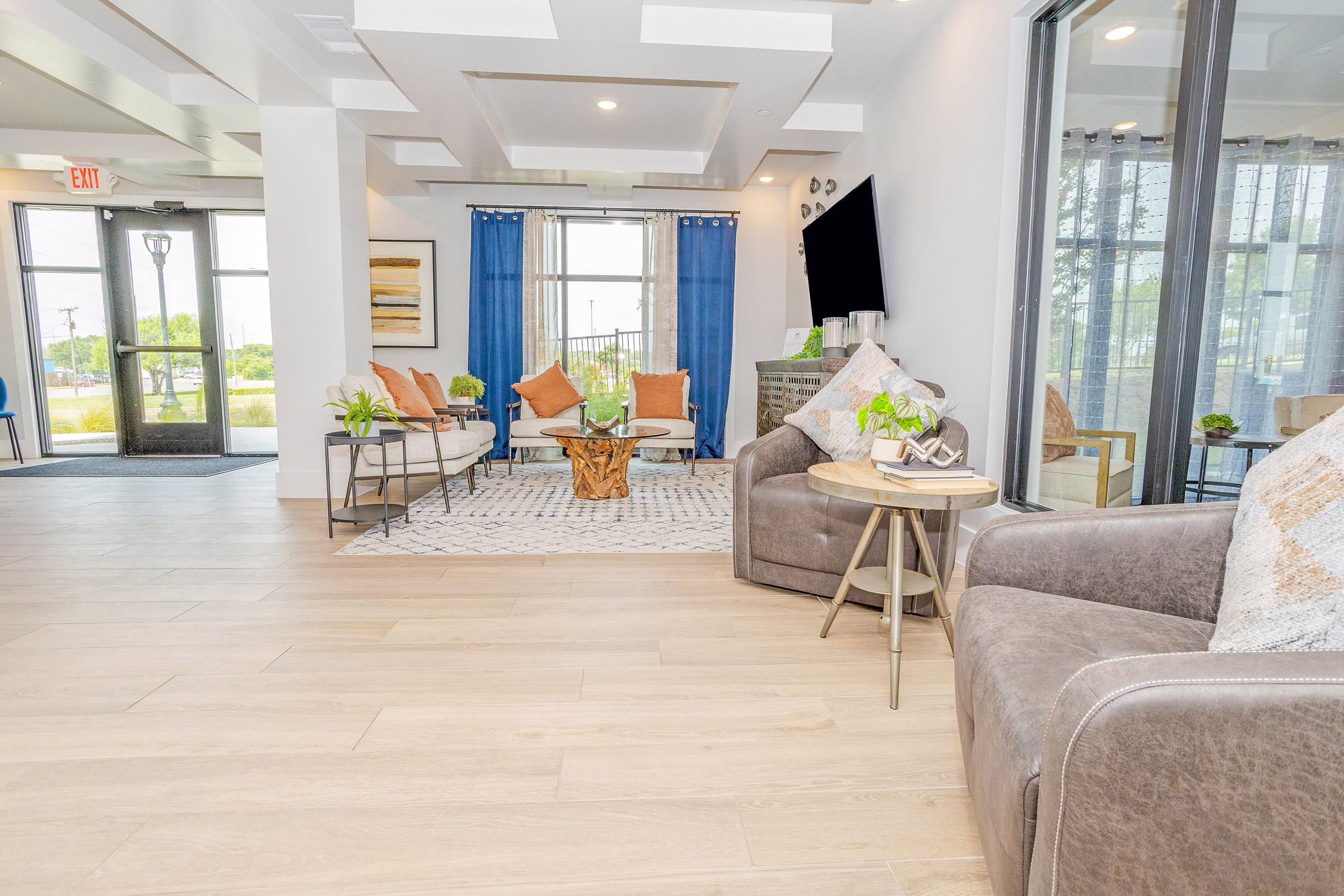A welcoming lobby area featuring two comfortable chairs, a coffee table made of wood, and a stylish rug. The space is decorated with vibrant cushions, potted plants, and a large window with blue curtains, allowing natural light to fill the room. A modern TV and artwork add to the contemporary design.