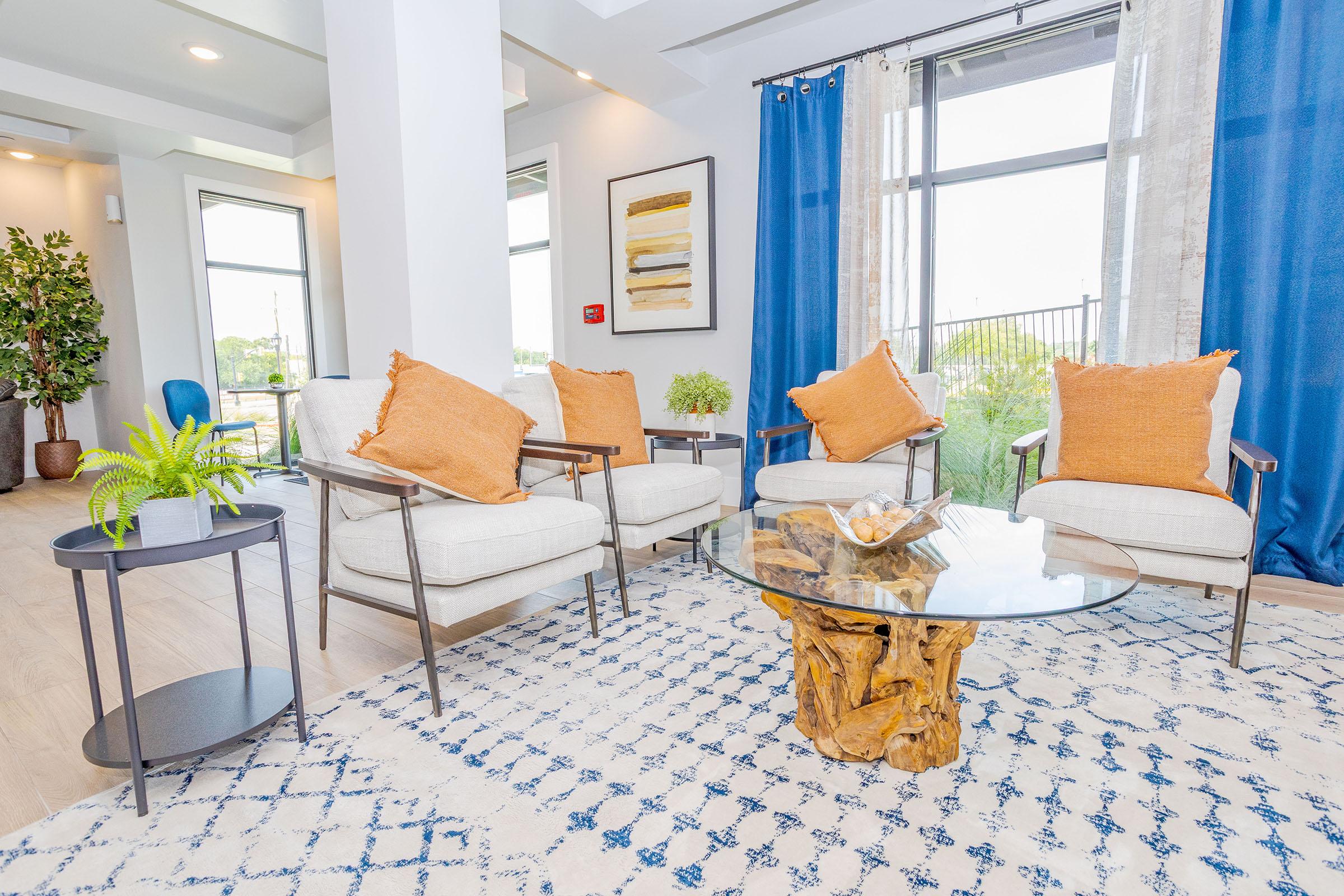 A modern living space featuring four white chairs with orange cushions arranged around a round glass table on a textured rug. There are decorative plants, a small side table, and large windows with blue curtains, allowing natural light to fill the room.