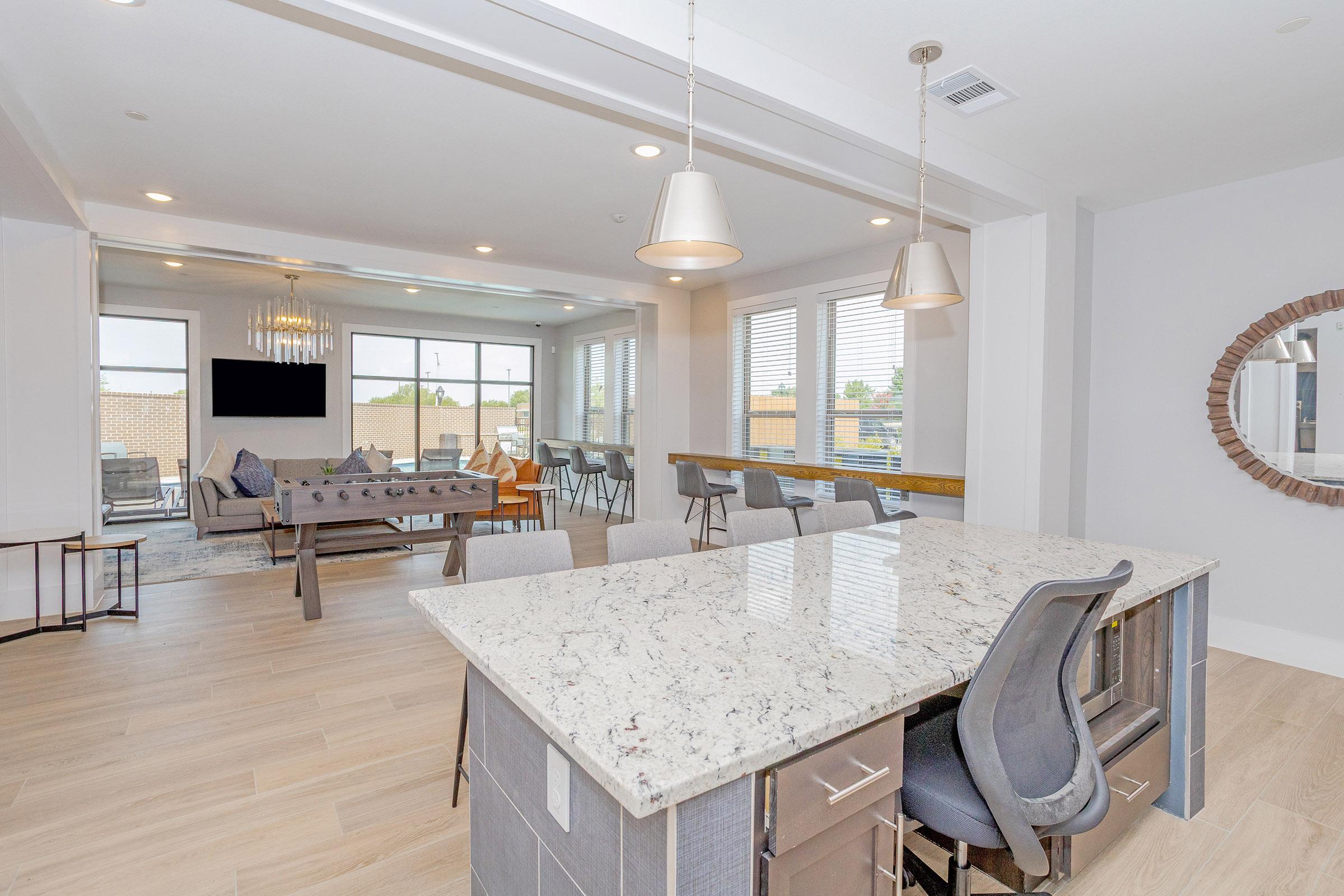 A bright, modern open-concept living space featuring a large kitchen island with granite countertops, pendant lighting, and bar stools. The area includes a cozy living room with large windows, a dining table, and stylish furnishings, creating a welcoming atmosphere.