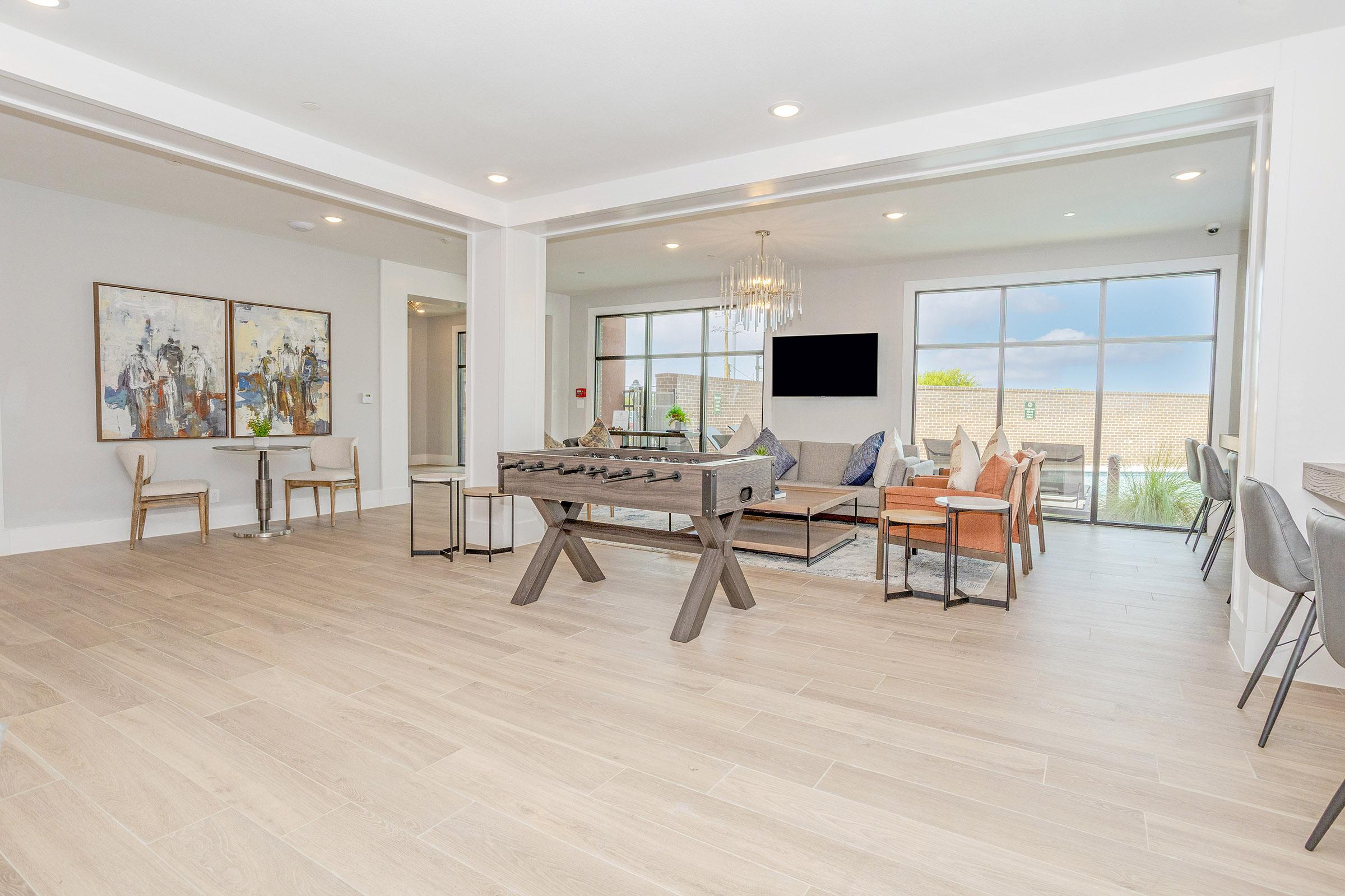 A modern living space featuring a foosball table, comfortable seating area with a sofa and armchairs, and a dining table. Large windows let in natural light, and the decor includes artwork on the walls. The flooring is light wood, creating a warm and inviting atmosphere.