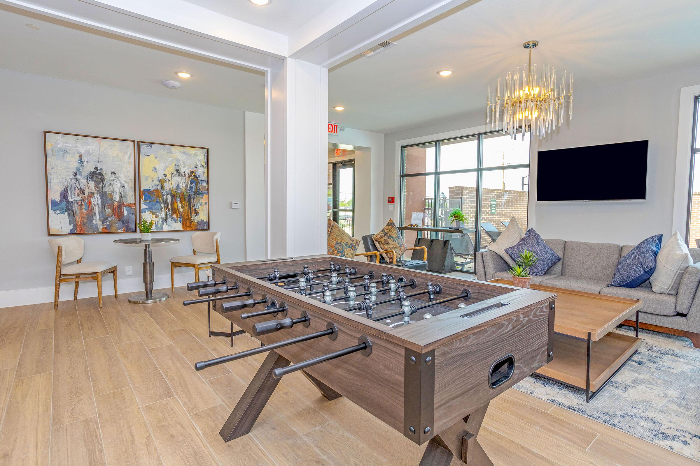 A modern lounge area featuring a foosball table in the foreground, a stylish sofa with blue and grey cushions, a light fixture overhead, and two pieces of abstract art on the walls. A table with two chairs is visible, along with a large window allowing natural light to fill the space.