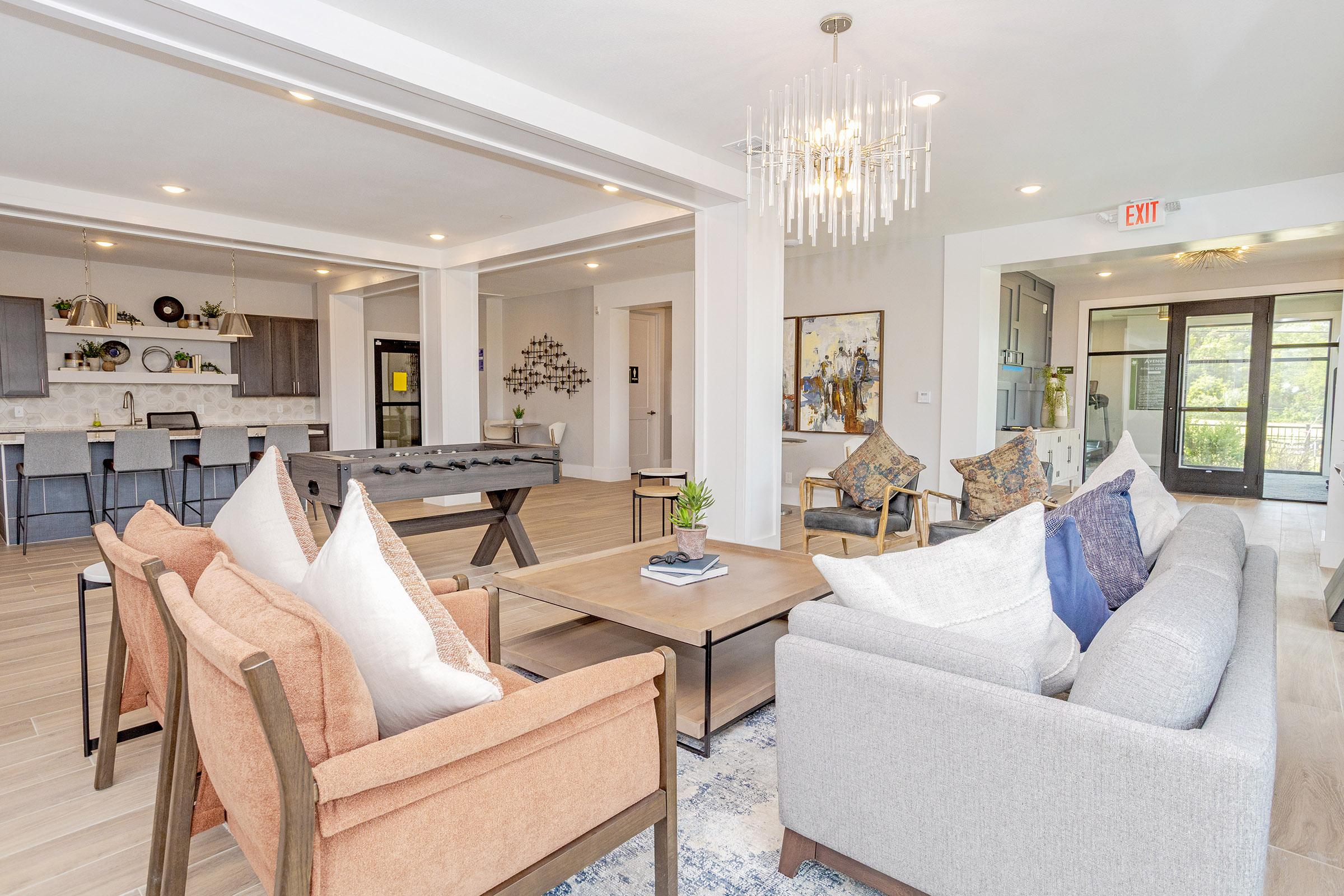 A modern living space featuring a cozy seating arrangement with a gray sofa and orange armchairs, a wooden coffee table, and decorative pillows. In the background, a foosball table is visible, along with a stylish kitchen area equipped with bar stools. Large windows allow natural light to fill the room, enhancing the inviting atmosphere.