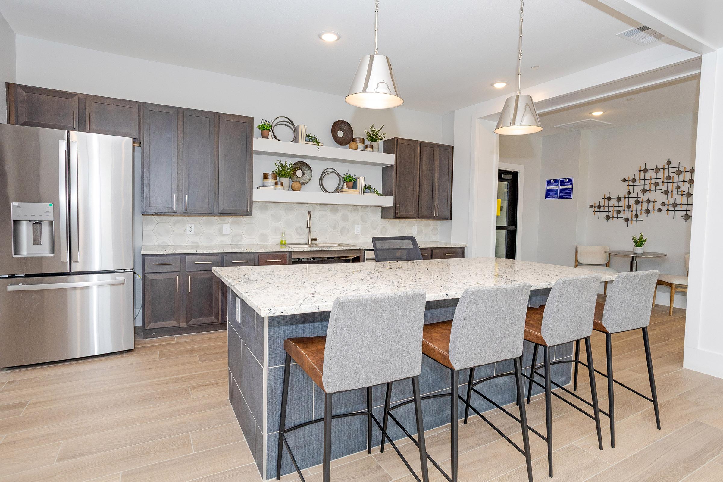 Modern kitchen featuring dark wood cabinets, a large granite island with seating for four, stainless steel appliances including a refrigerator, and stylish pendant lighting. The backsplash has a neutral tile pattern, and decorative plants are placed on shelves. The room is bright and contemporary.