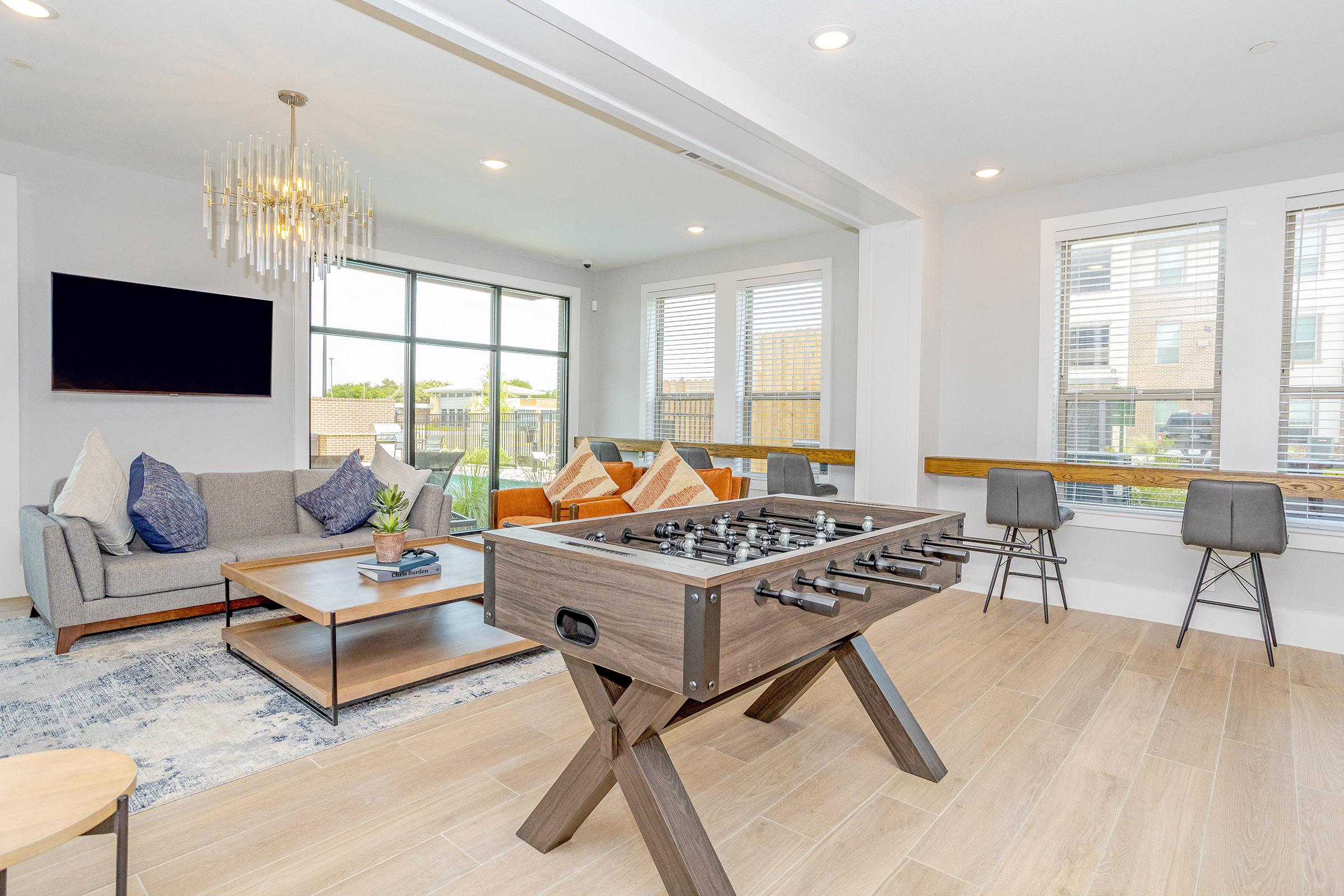 A modern living room featuring a foosball table in the foreground, a cozy gray sofa with decorative pillows, a wooden coffee table, and large windows letting in natural light. The decor includes a chandelier and indoor plants, with stylish seating options visible in the background.