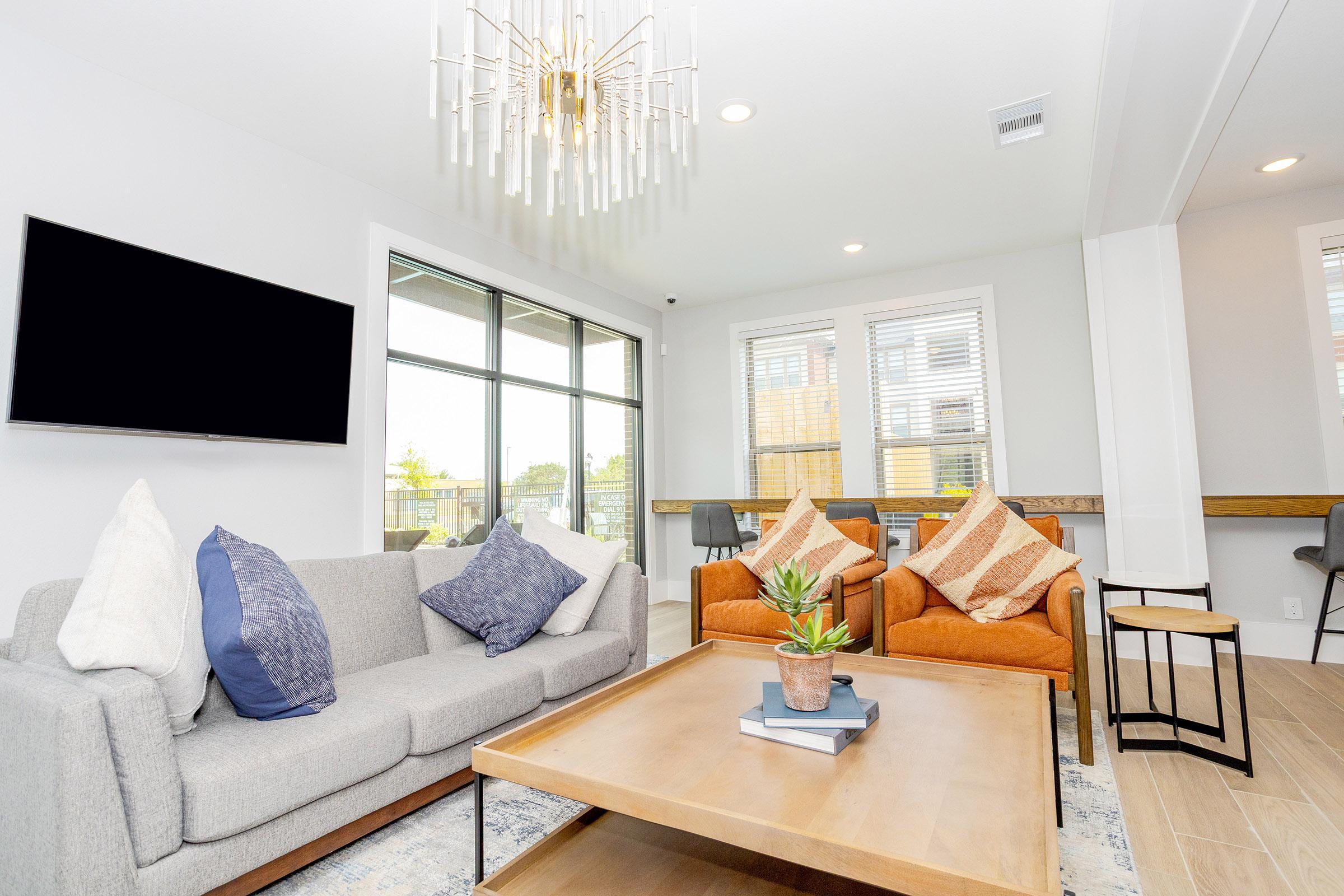 A modern living room featuring a gray sofa with decorative pillows, two orange armchairs, a wooden coffee table with a potted plant, and a wall-mounted TV. Large windows allow natural light to fill the space, complemented by a stylish chandelier overhead and a workspace in the background.