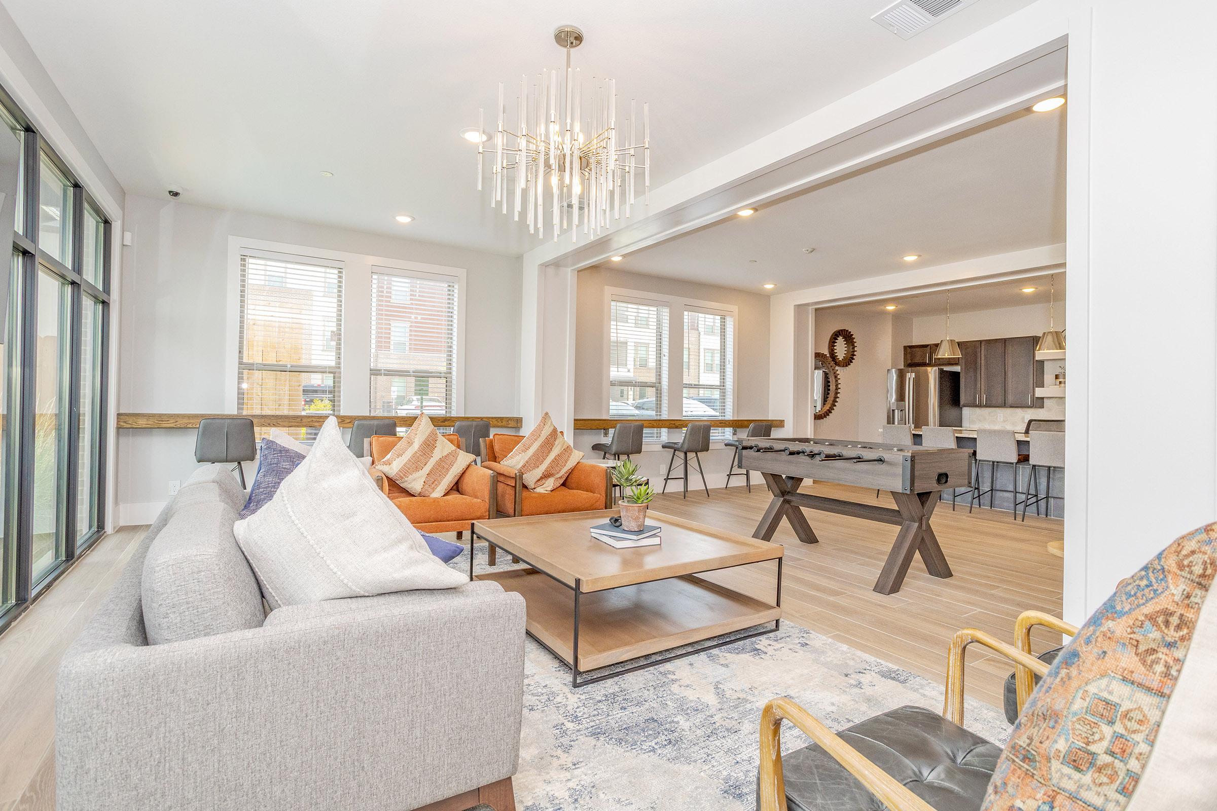 A modern, inviting living space featuring a gray sofa with patterned pillows, an orange armchair, and a wooden coffee table. A foosball table is visible in the background, along with a bright kitchen area and large windows letting in natural light. Stylish light fixtures and decor add to the contemporary feel.