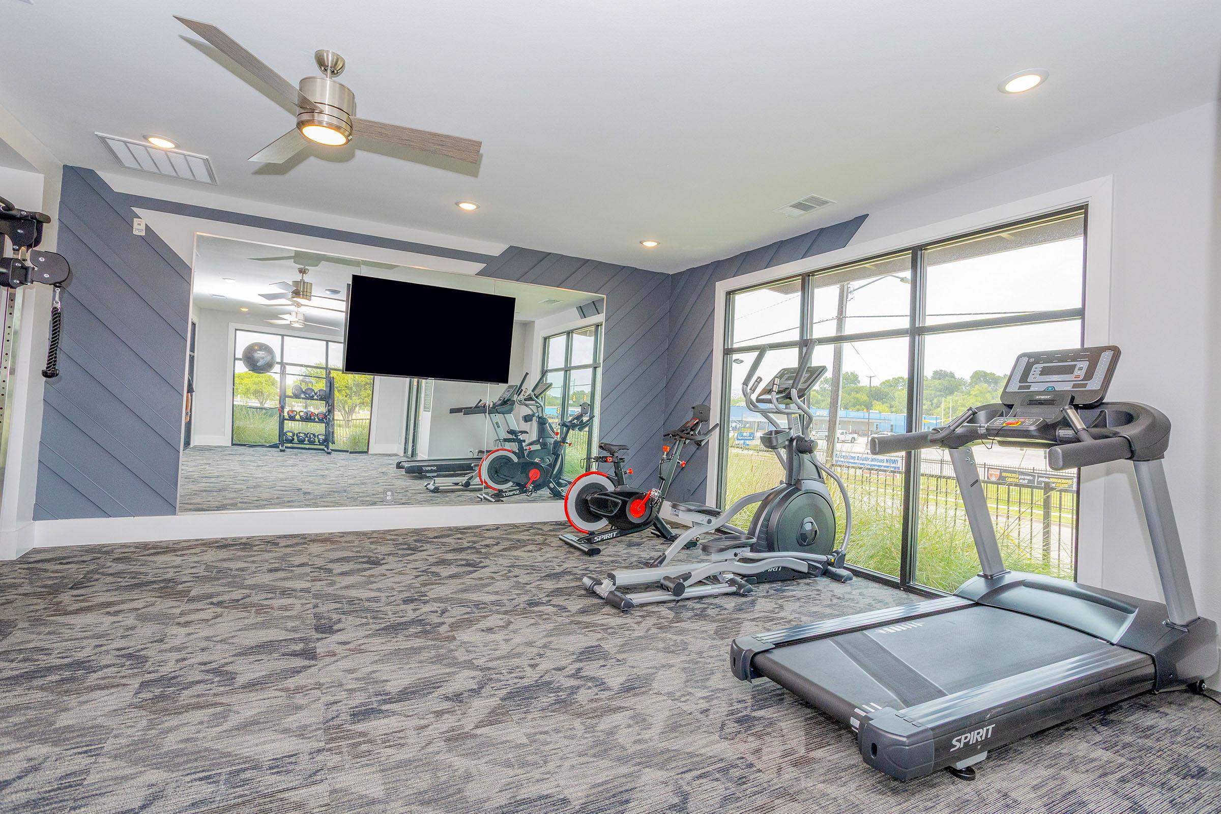 A modern fitness room featuring a treadmill, an elliptical machine, and a large flat-screen TV. The walls are painted in a stylish design, and there are large windows showing a view outside. The flooring is carpeted, creating a comfortable workout environment.
