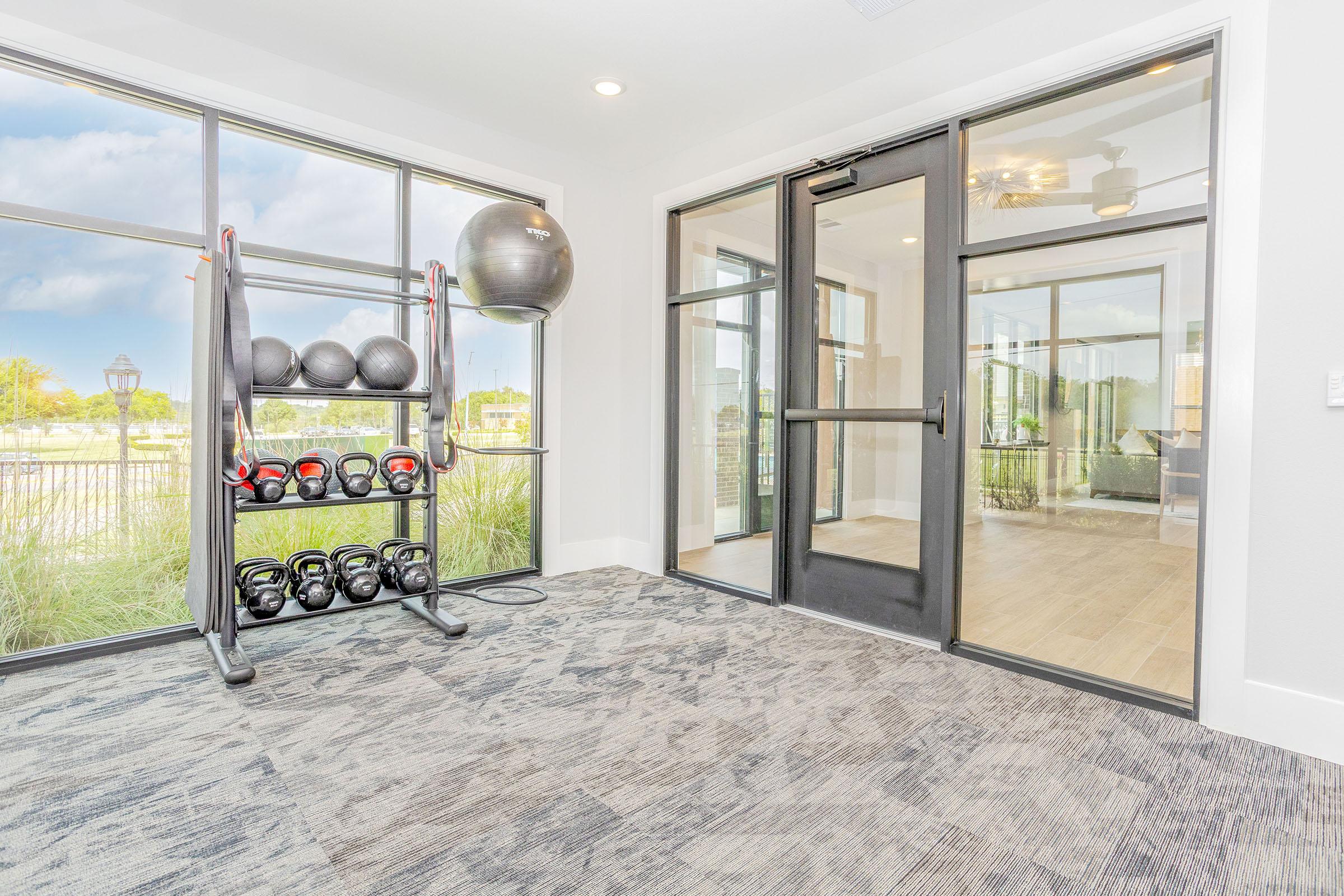 A modern gym area featuring a variety of fitness equipment, including weights and exercise balls. Large windows allow natural light, and the space has a clean, contemporary design with a neutral color palette and a carpeted floor. A glass door leads to another area.
