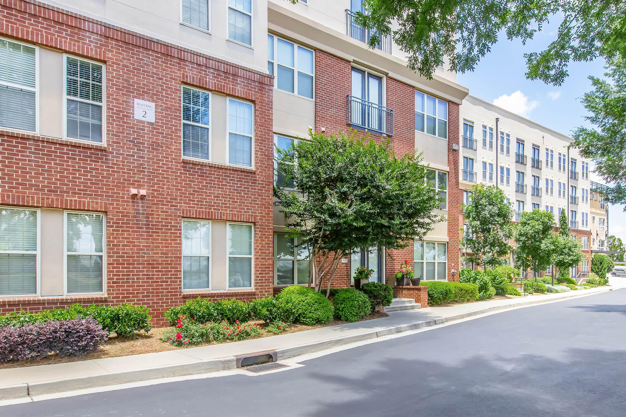 A modern apartment building with a mix of red brick and beige facade. The exterior features large windows, greenery, and landscaped bushes along a smooth asphalt road. Bright sunlight enhances the inviting atmosphere of the residential area.