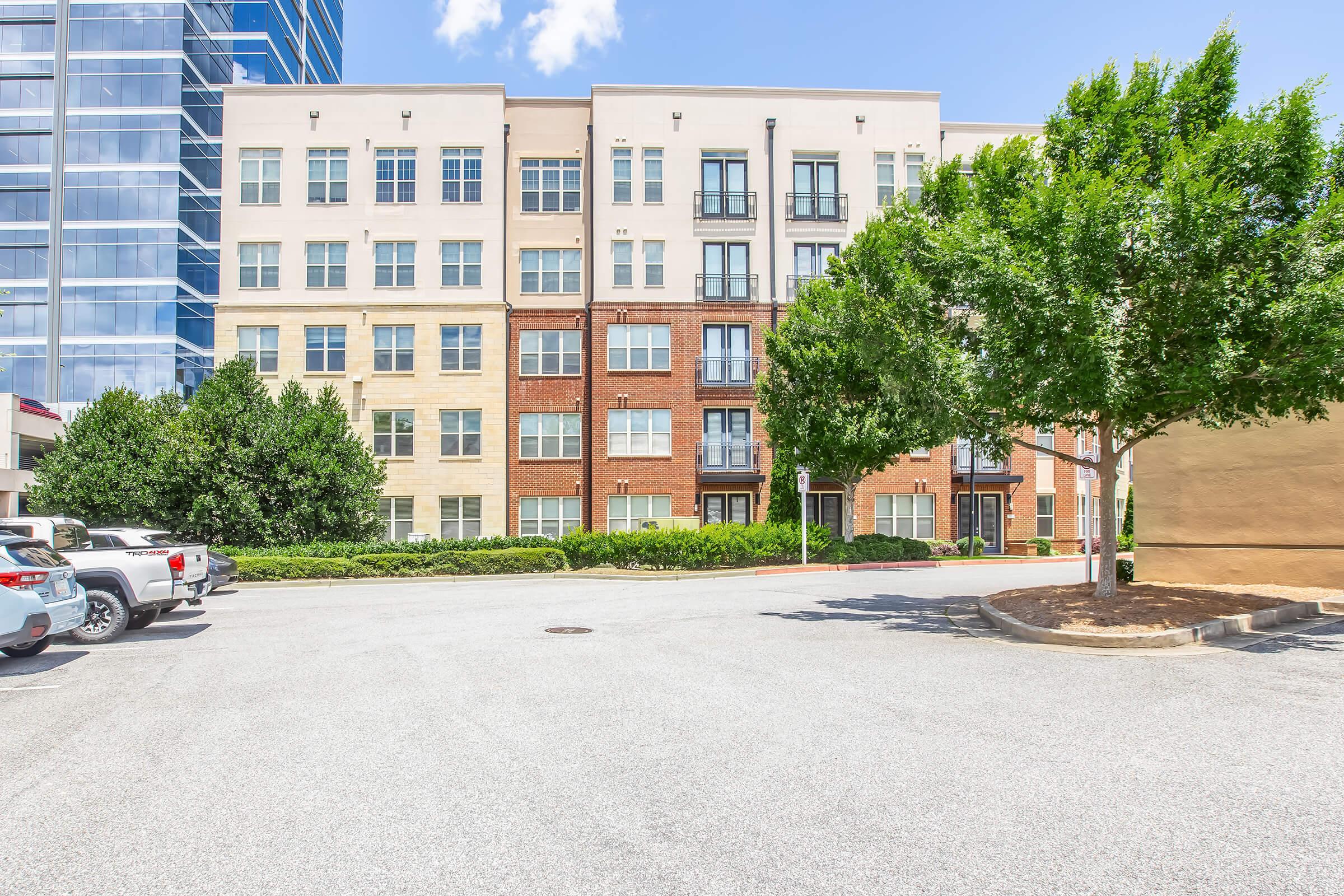 Modern apartment building with a mix of beige and brick exteriors. Surrounded by lush greenery, it features large windows and balconies. A clear blue sky is visible overhead, and there is a parking area in the foreground.