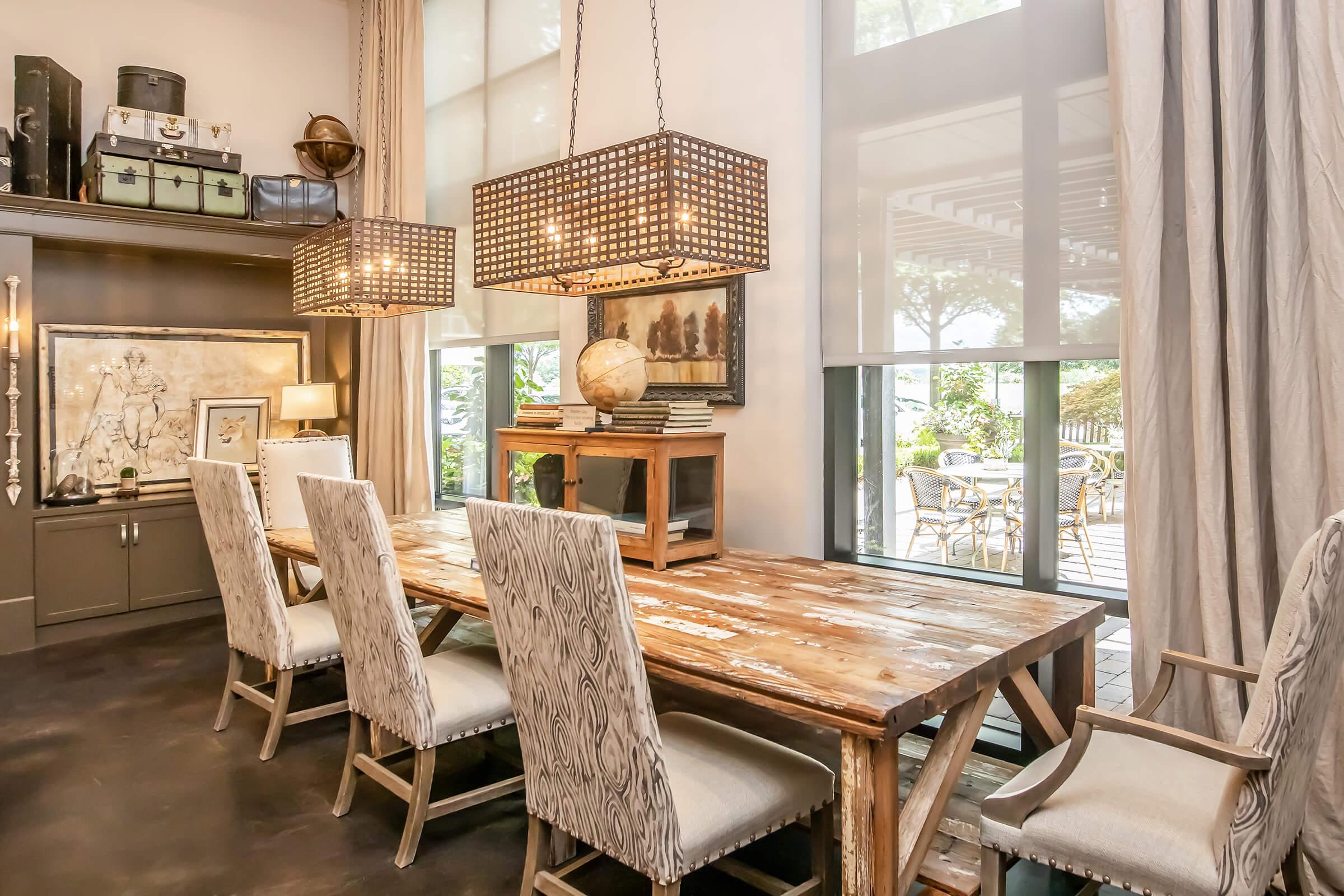 A stylish dining area featuring a large wooden table surrounded by upholstered chairs. Two modern pendant lights hang above the table. In the background, large windows provide a view of an outdoor patio with seating. Shelves hold decorative items and a globe adds a touch of sophistication to the space.