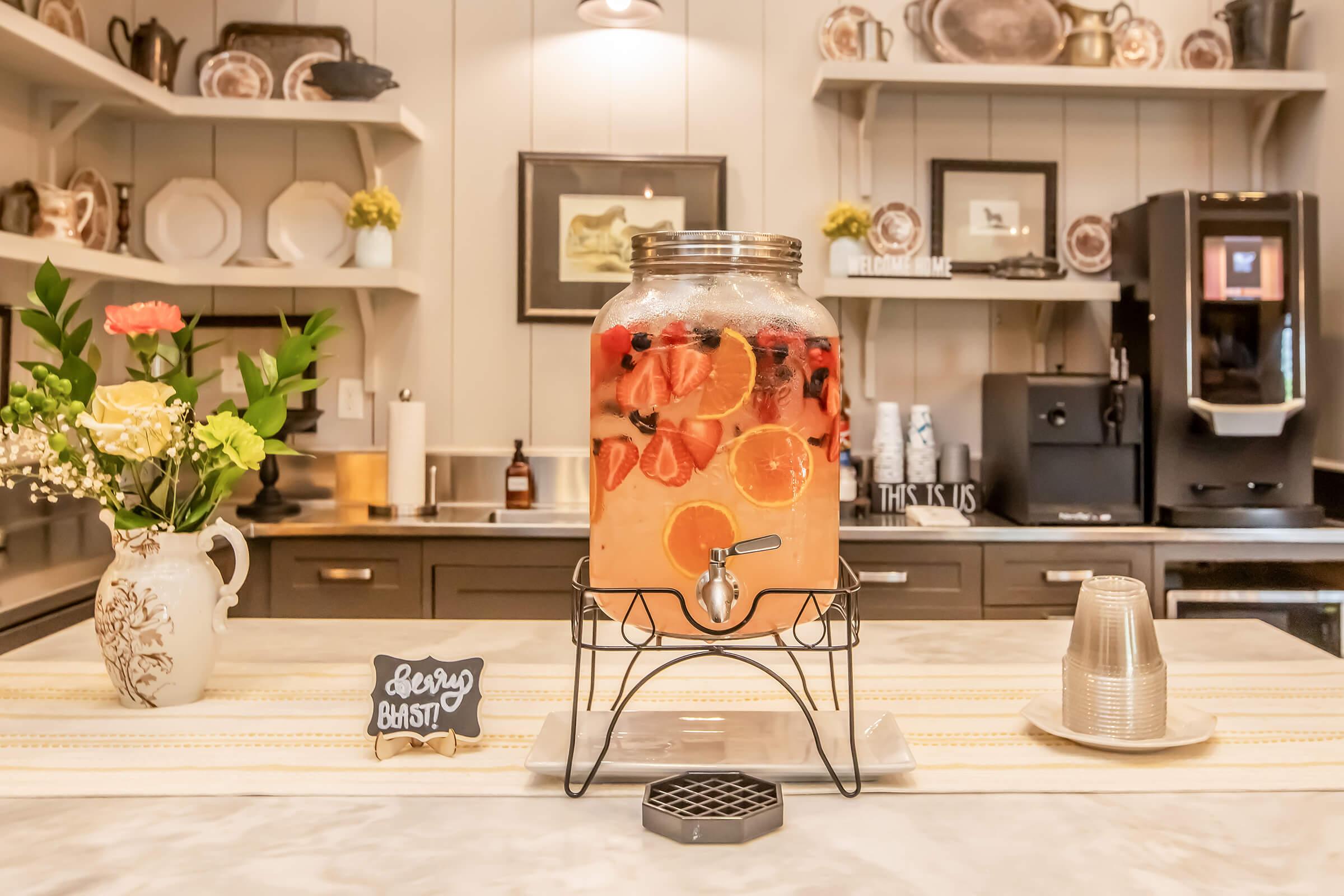 A large glass beverage dispenser filled with colorful fruit-infused drink, featuring slices of orange, strawberries, and blueberries. It is placed on a marble countertop in a cozy kitchen, alongside a small chalkboard sign and a vase of flowers, with vintage dishware on the shelves in the background.