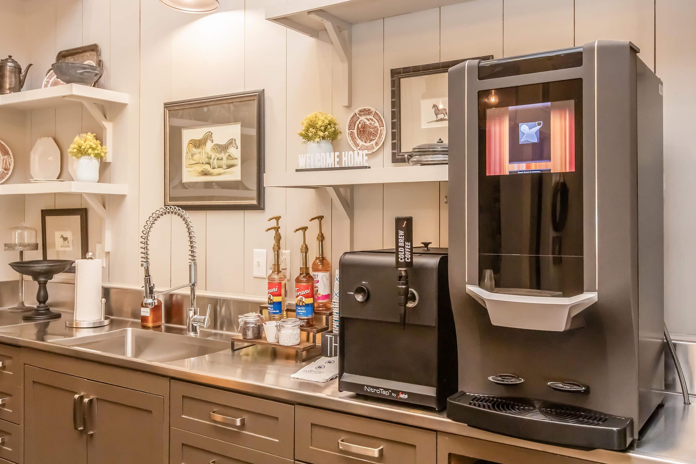 A modern kitchen setup featuring a stainless steel countertop with an ice maker and drink dispenser. Shelves above hold decorative items and bottles. The ambiance is warm and inviting, with framed pictures and small plants on display.