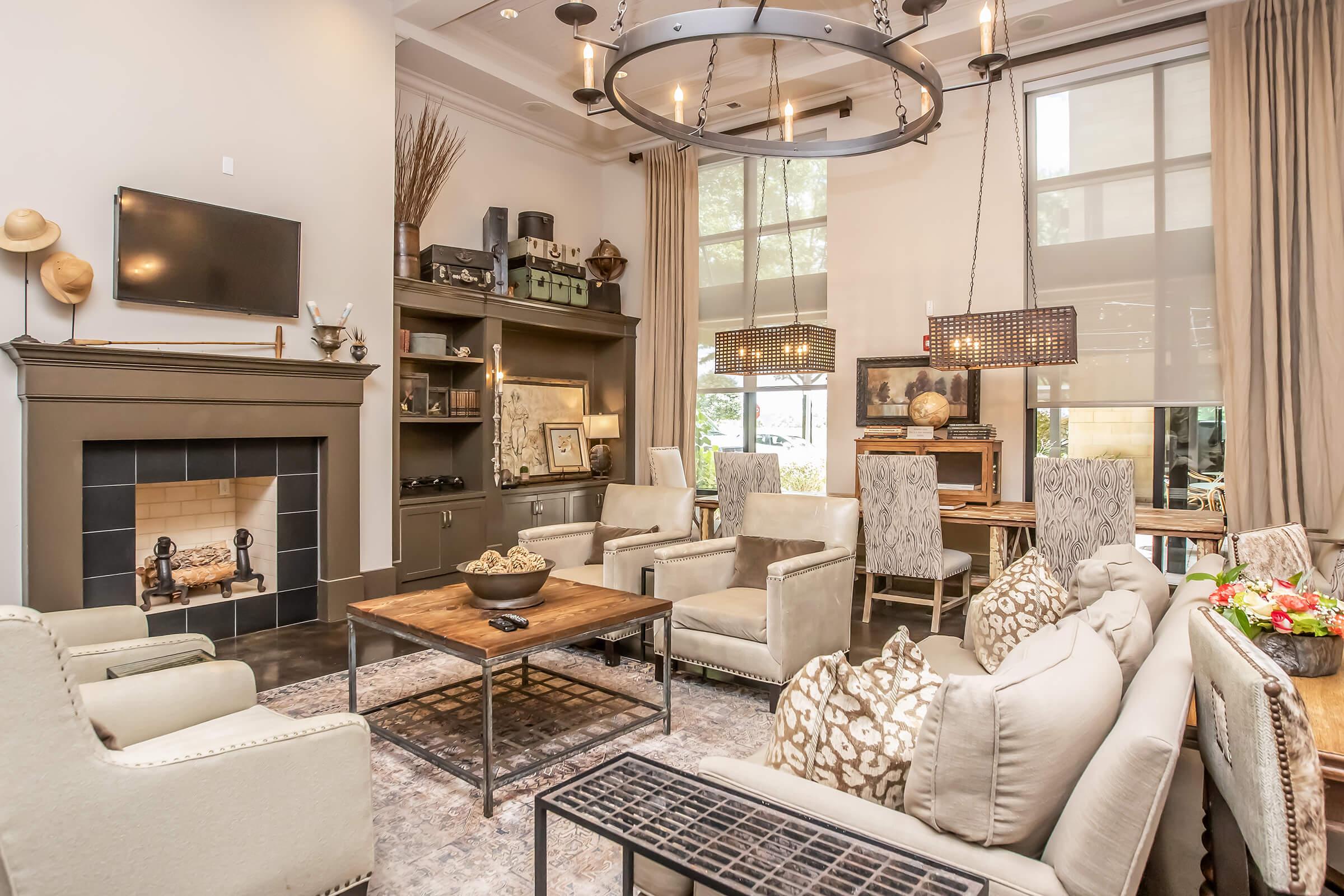 A cozy, modern living room featuring a fireplace, large windows with flowing curtains, and a mix of comfortable seating. The decor includes a coffee table with decorative elements, a stylish chandelier, and tasteful wall art. The space is warmly lit and inviting, ideal for gatherings or relaxation.