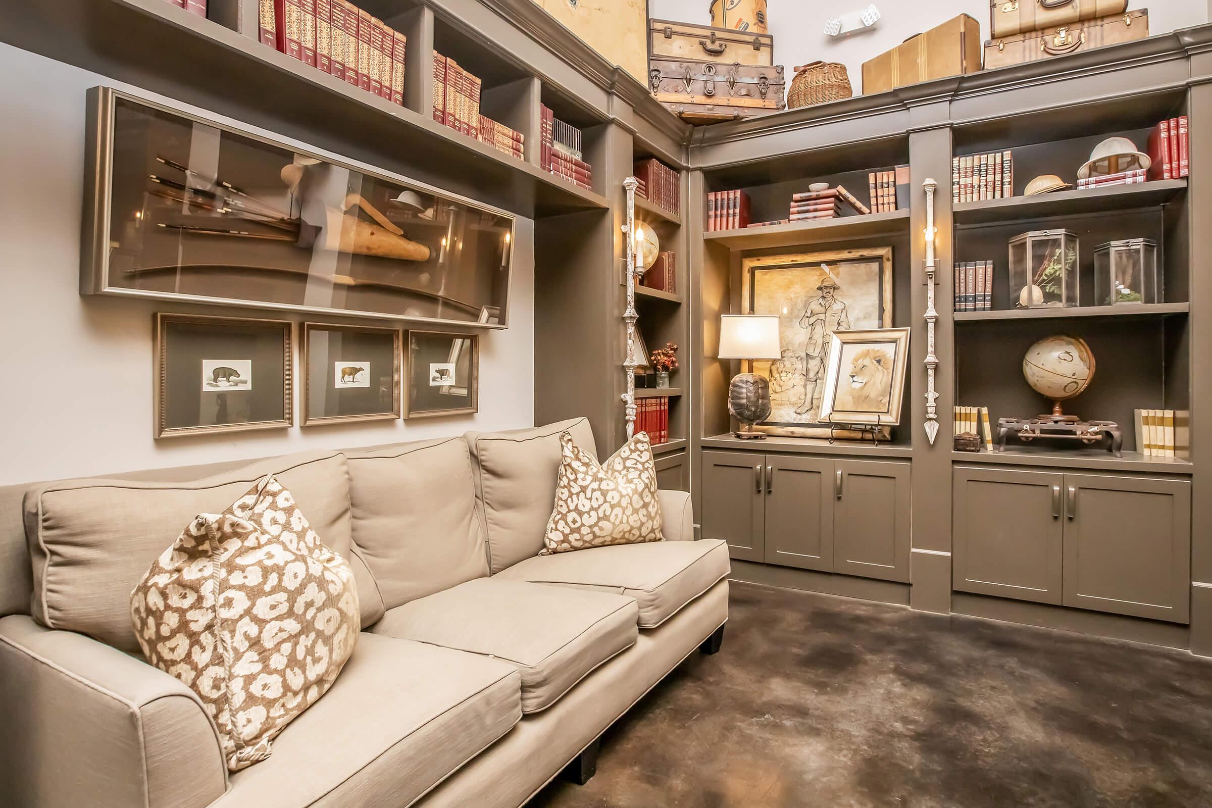 A cozy, stylish reading nook featuring a comfortable beige sofa with patterned throw pillows. Behind it is a gray bookshelf filled with books, decorative items, and artwork, including a vintage airplane display. Warm lighting enhances the inviting atmosphere of the space.