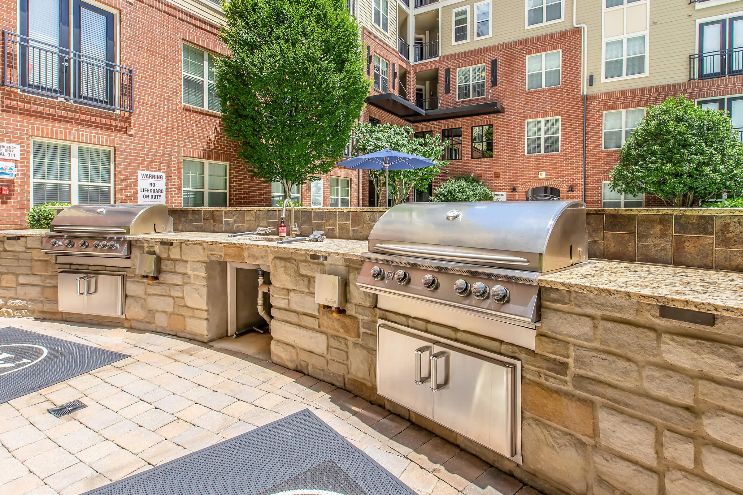Outdoor grilling area featuring two stainless steel gas grills set against a stone counter, surrounded by lush greenery. The space is designed for social gatherings, complete with paved flooring and an umbrella for shade, located in a residential complex.