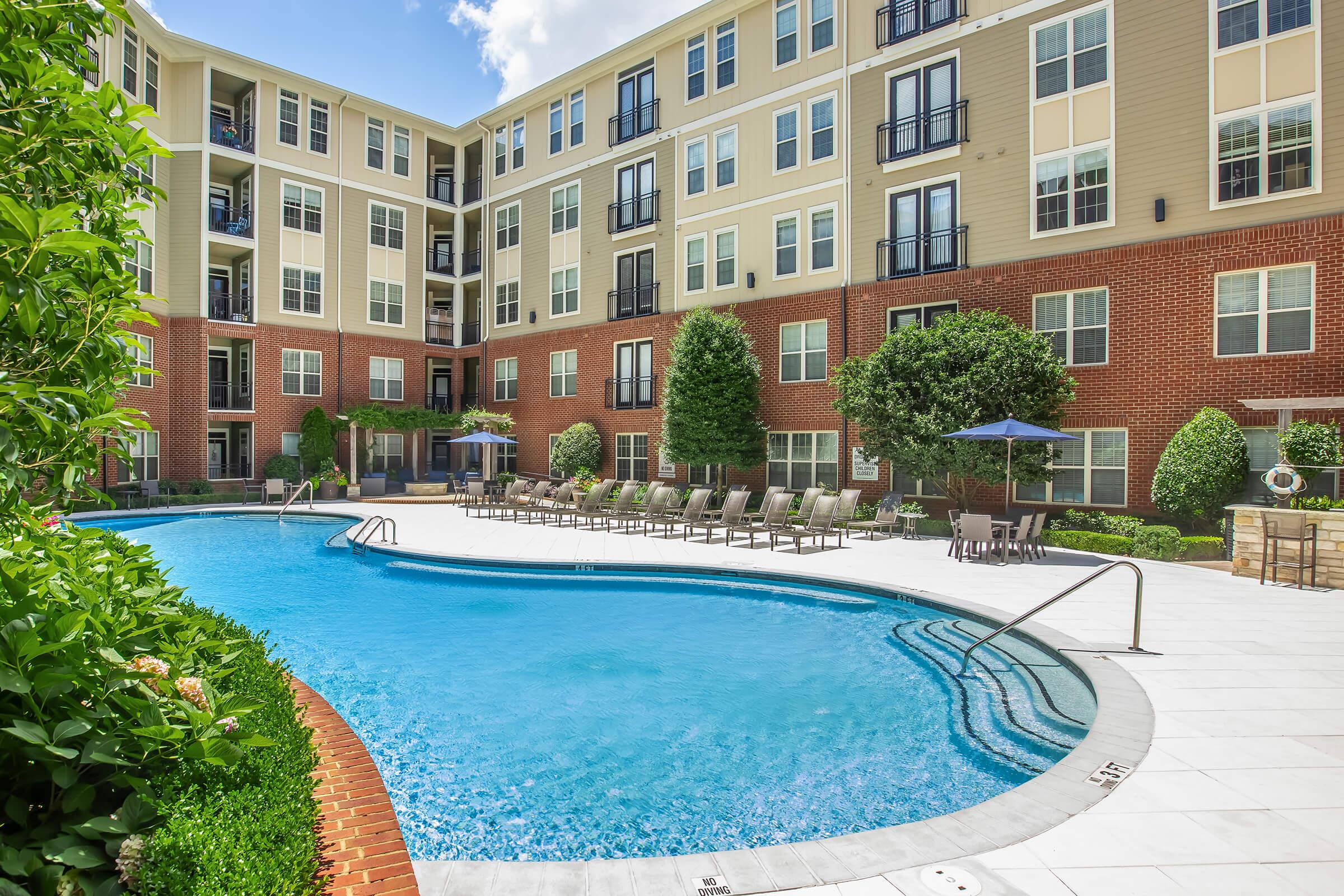 A well-maintained outdoor swimming pool surrounded by landscaped areas and residential buildings. Lounge chairs are neatly arranged around the pool, and shade umbrellas are placed for comfort. The setting is bright and inviting, suggesting a relaxation space in an apartment complex.