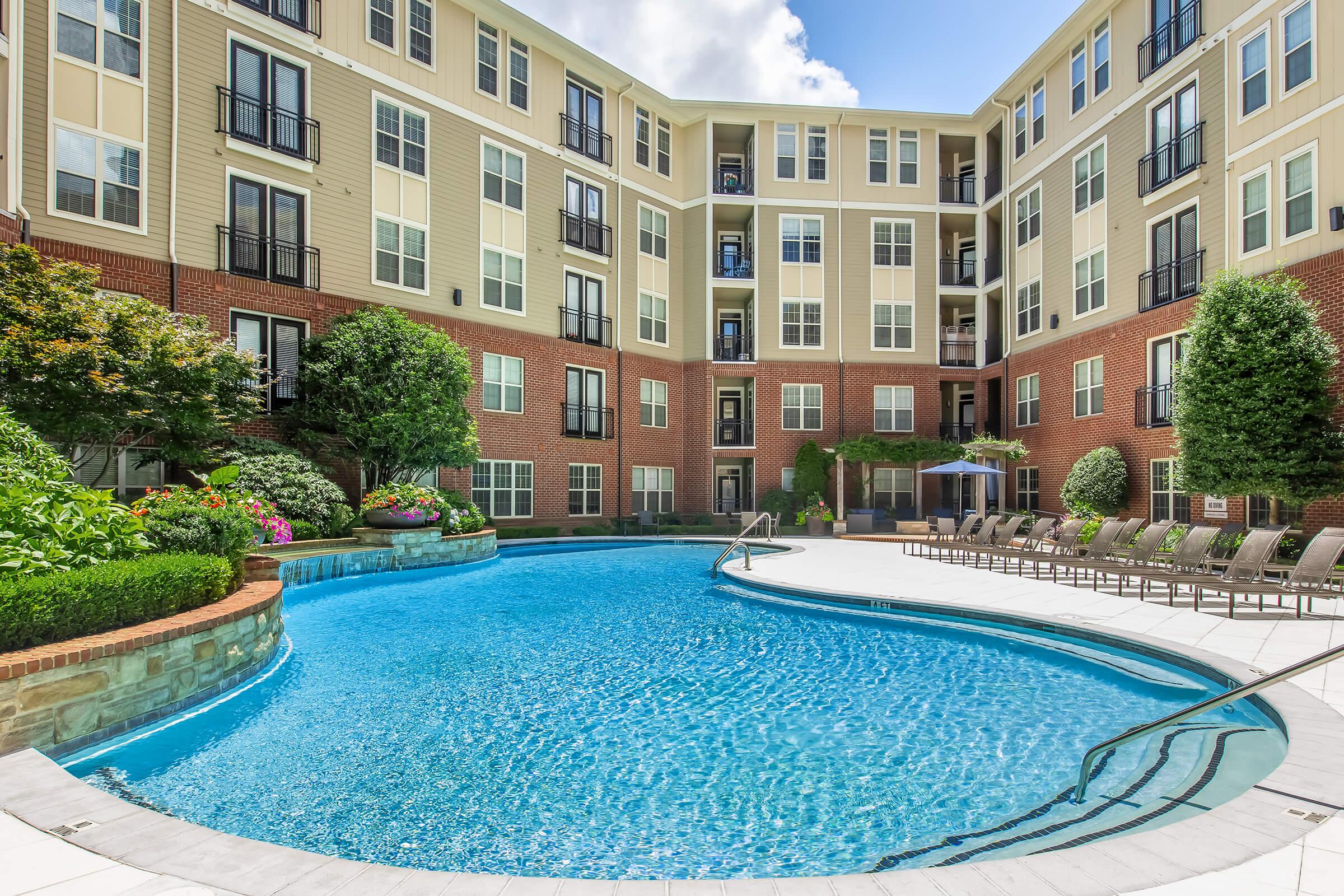 A bright, inviting outdoor pool surrounded by modern apartment buildings. The pool features a curved edge with lounge chairs arranged nearby. Lush greenery and flowerbeds provide a colorful backdrop, while umbrellas offer shade. The overall scene conveys a relaxing, resort-like atmosphere.